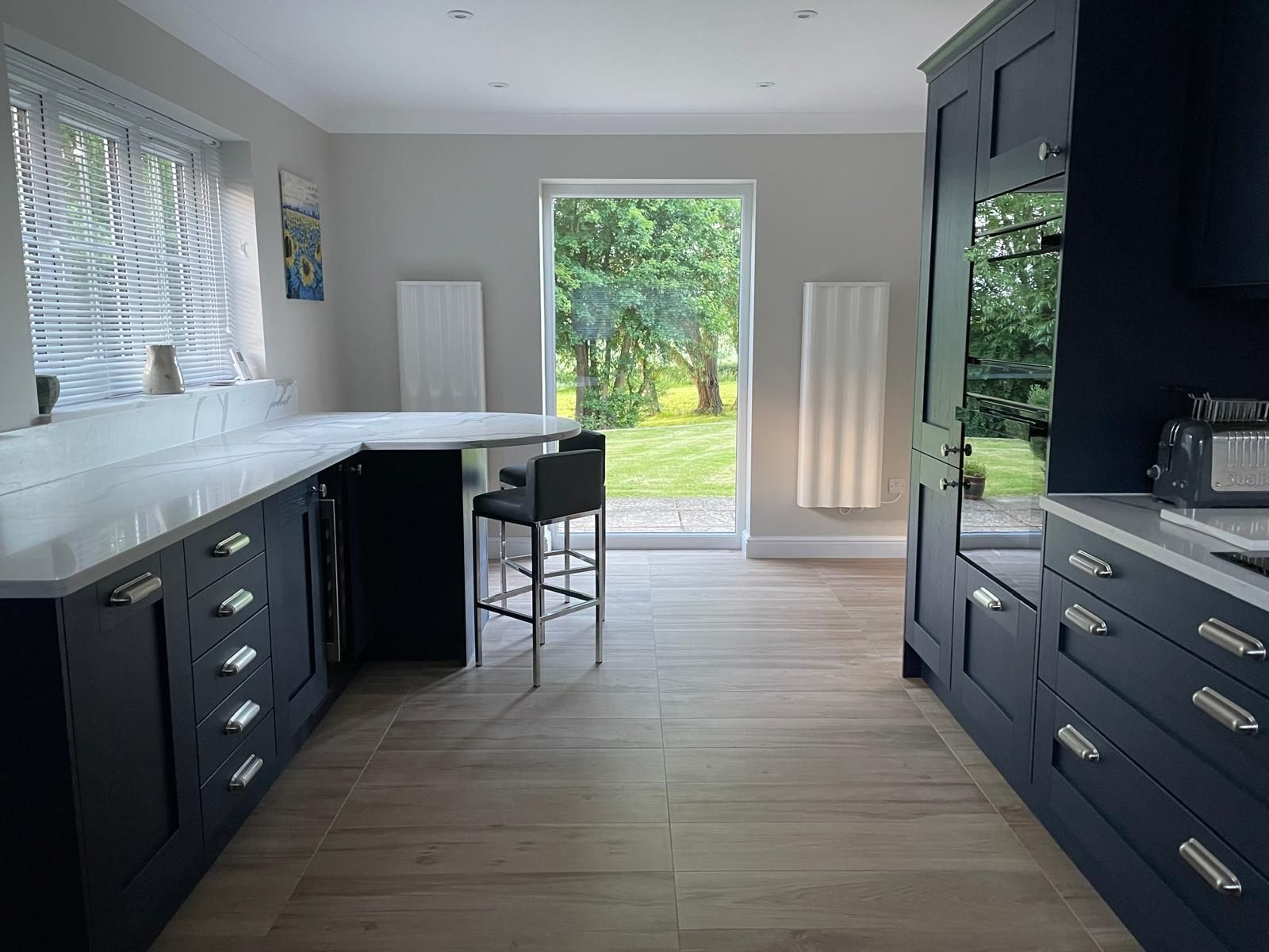 A modern kitchen with dark blue cabinets, light wood floors, a breakfast bar, and a doorway leading to a grassy garden.