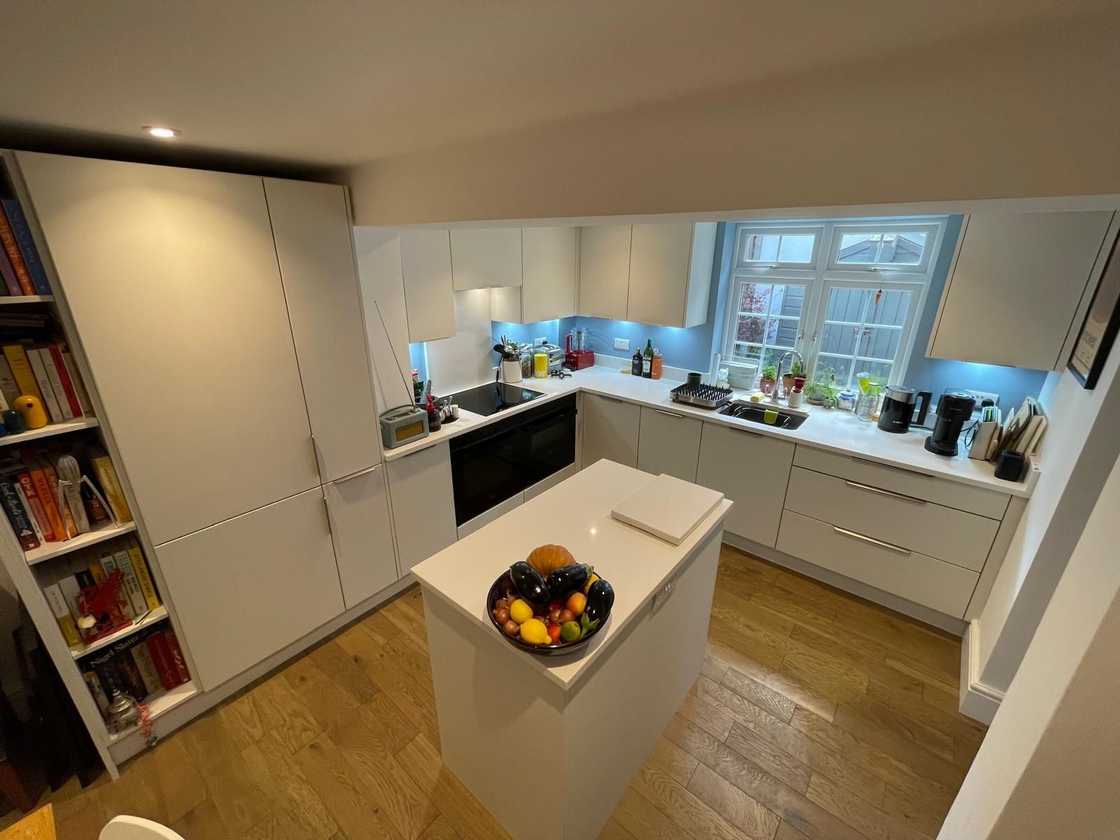A modern white kitchen with an island, light wood flooring, a window above the sink, and a tall bookshelf on the left.