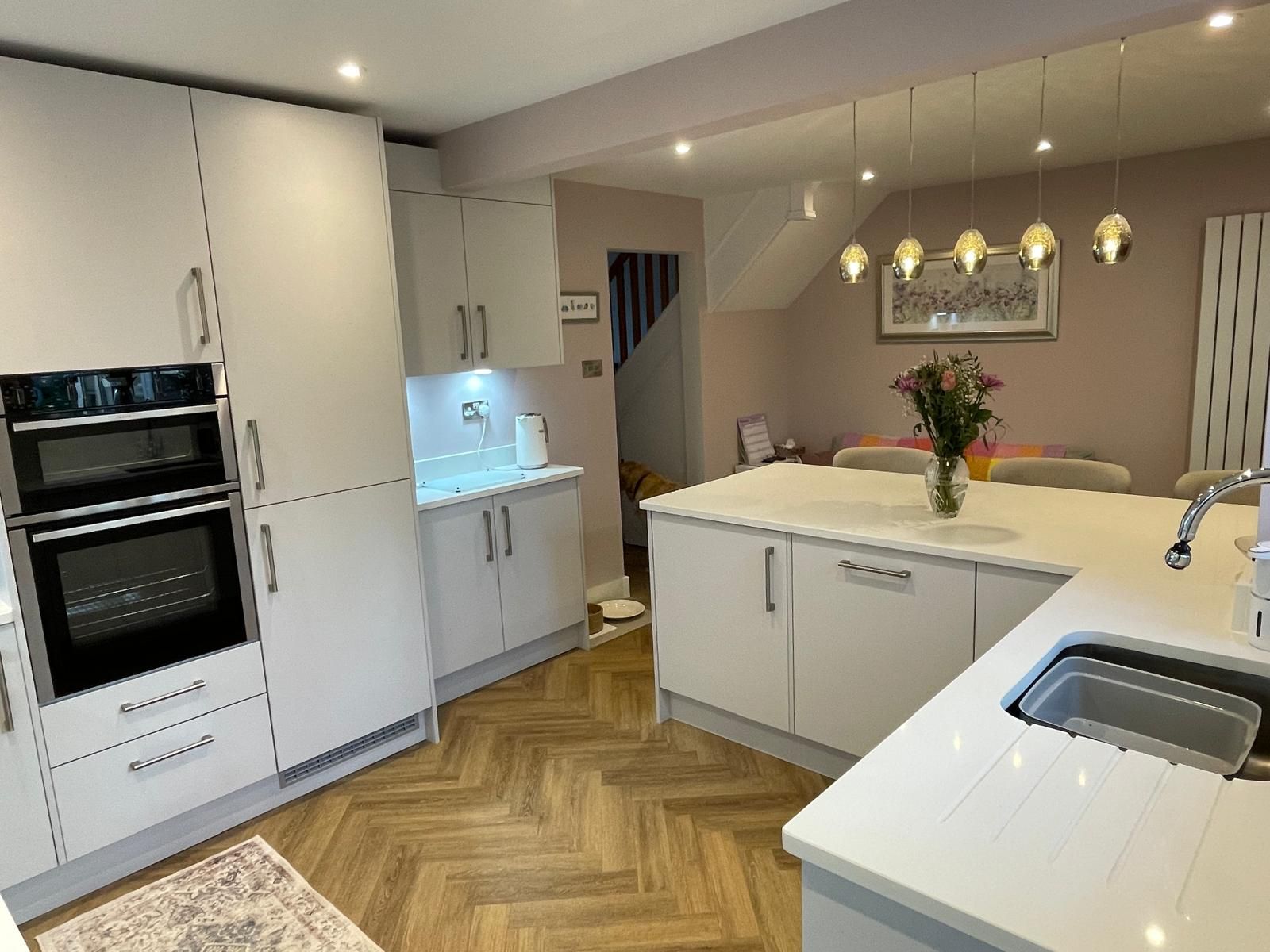 Modern white kitchen with herringbone wood floors, a large central island, double wall ovens, and pendant lighting.
