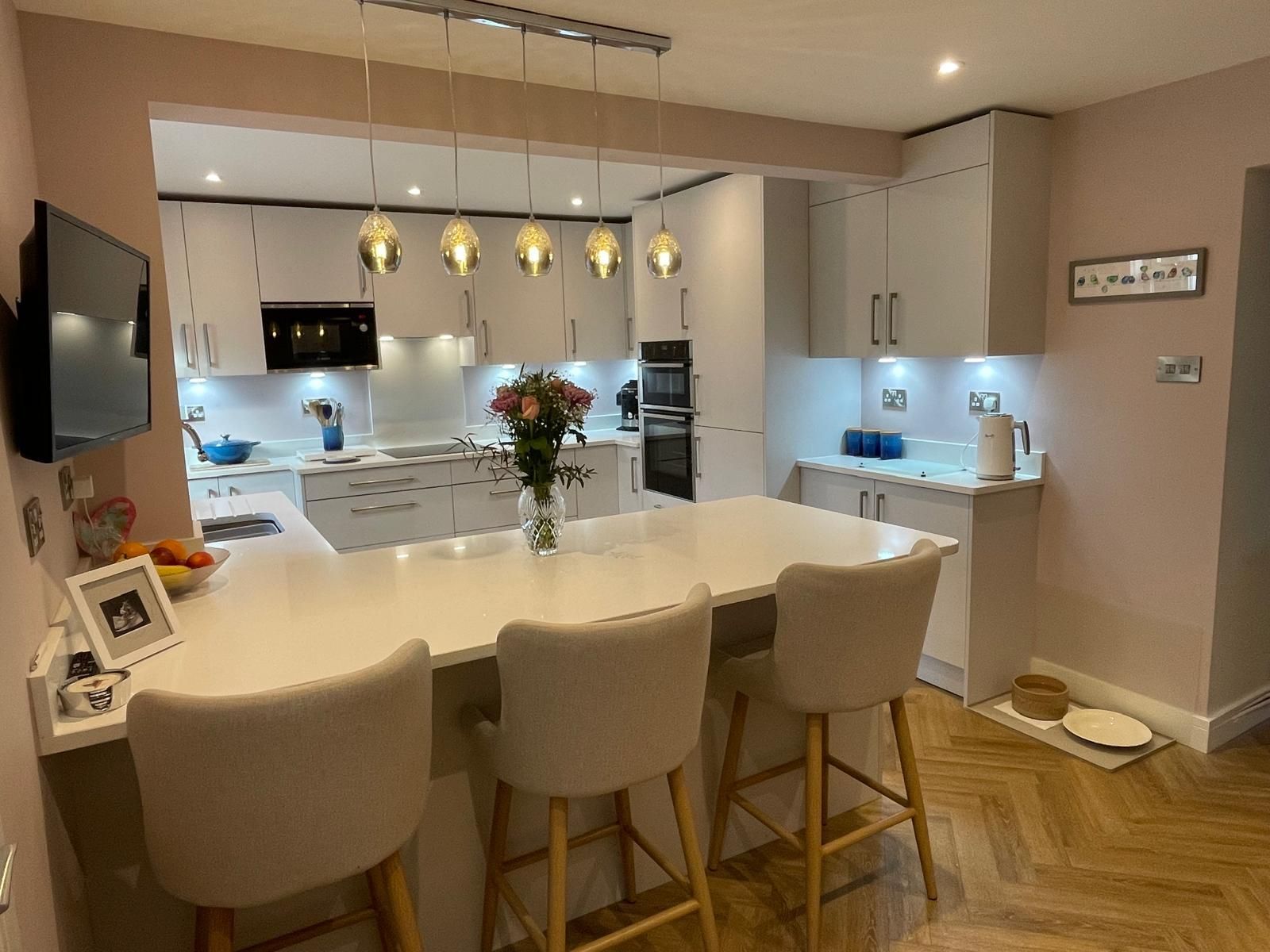 A modern, open-concept kitchen with white cabinets, a central island with three bar stools, and herringbone flooring.
