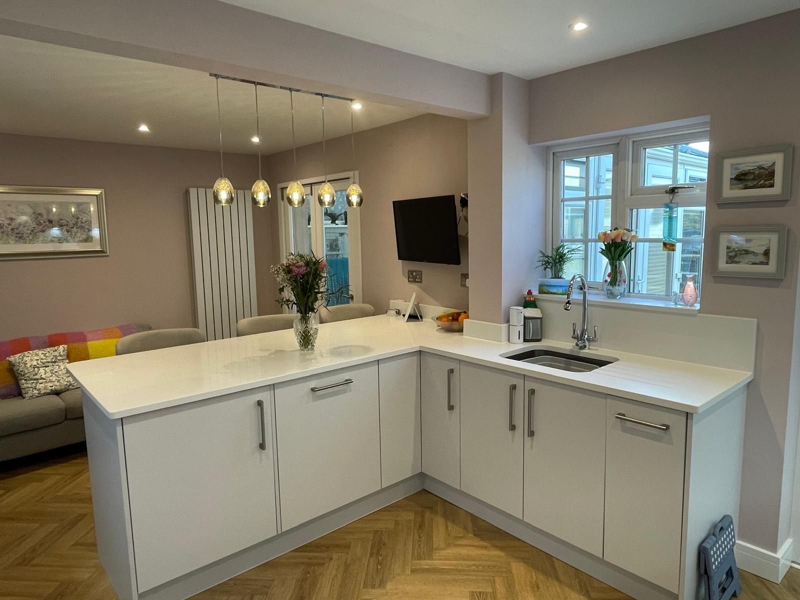 Modern kitchen featuring white L-shaped cabinets, a sink, a vase of flowers, and a dining area in the background.