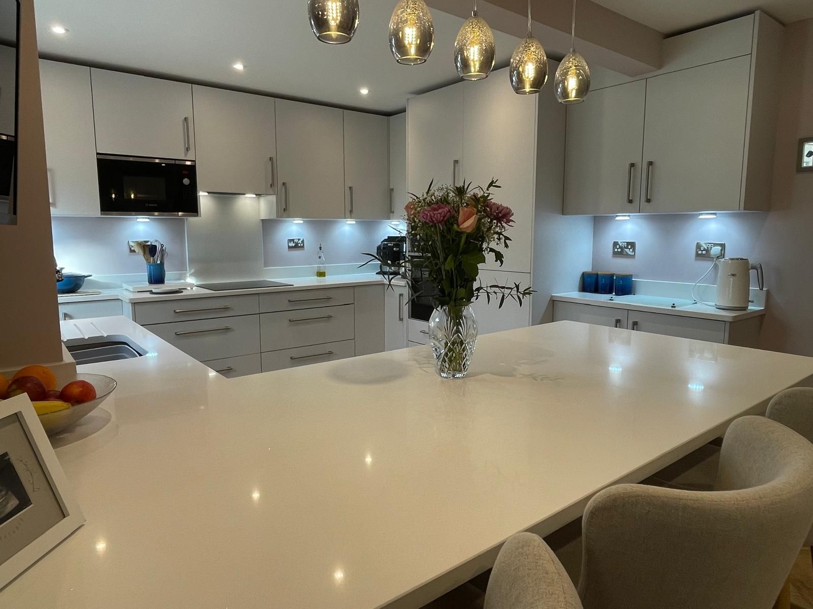 A modern kitchen with white cabinets, light countertops, an island with a flower vase, and hanging pendant lights.