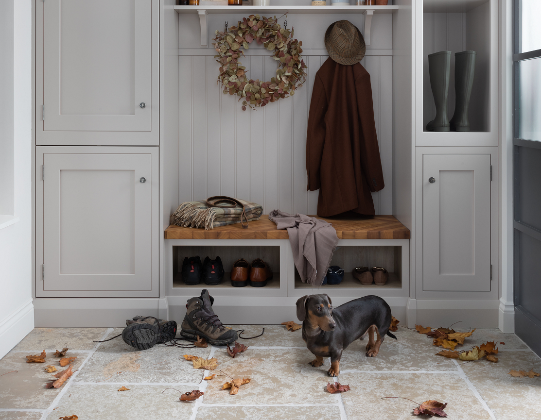 A dachshund stands on a stone floor with scattered autumn leaves in a mudroom featuring a bench, coat rack, and cabinets.