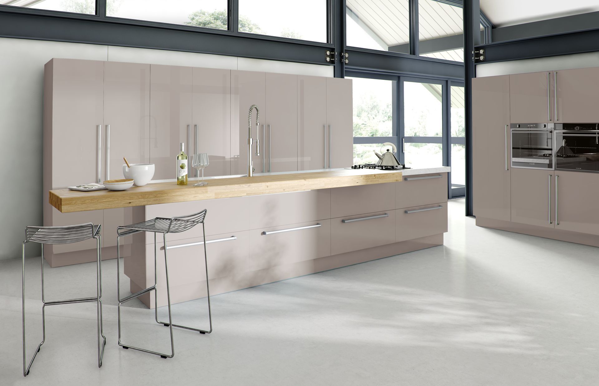 A modern kitchen with glossy taupe cabinets, a long natural wood breakfast bar, two metal stools, and large windows.