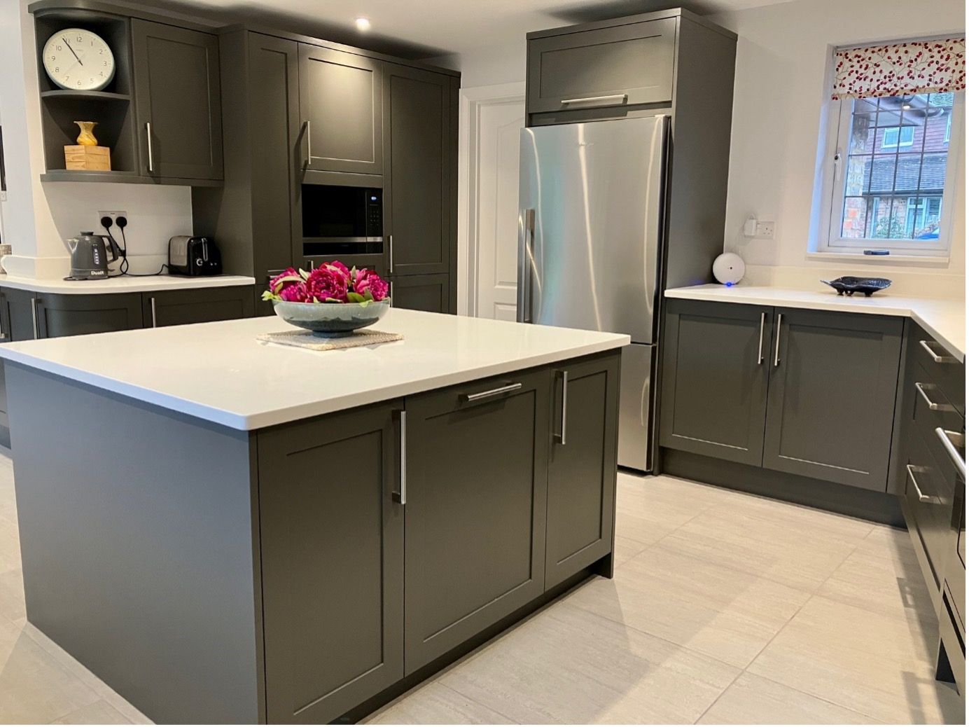 A modern kitchen featuring charcoal grey cabinets, white countertops, a central island, and a stainless steel refrigerator.
