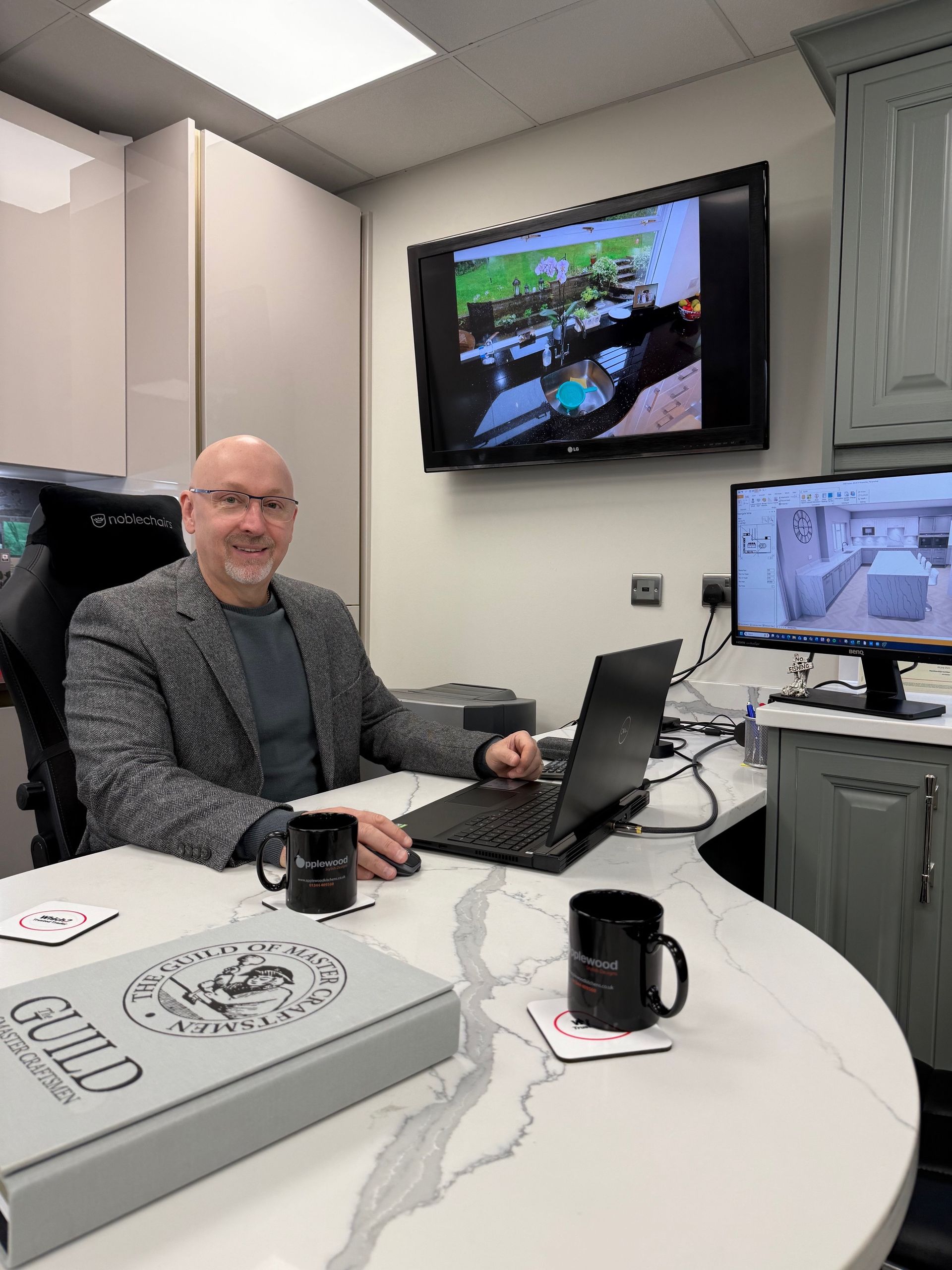A smiling man in a blazer sits at a desk with a laptop, monitors, and two mugs in a professional office setting.