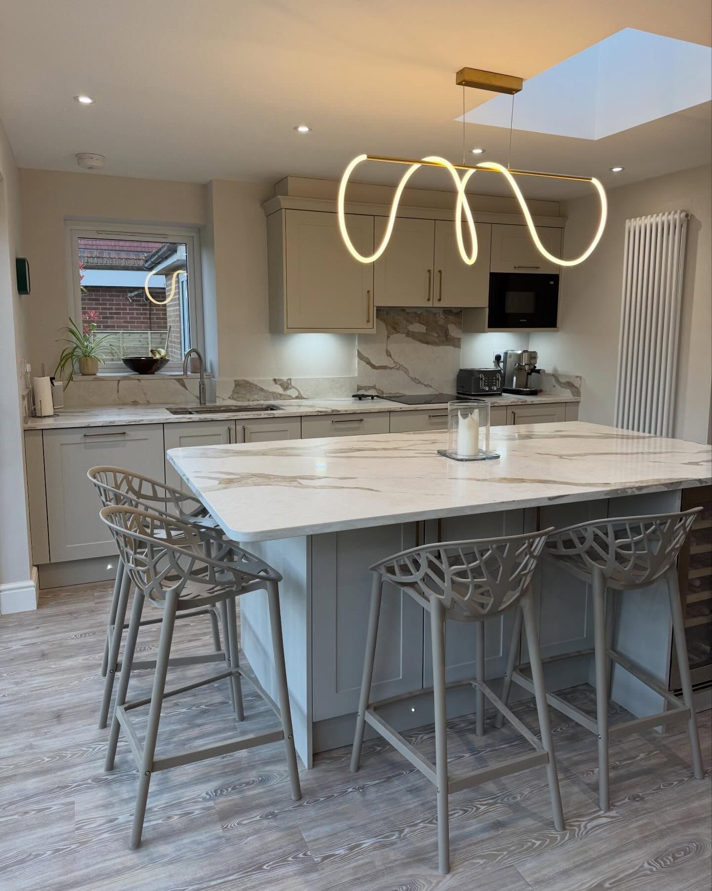 A modern kitchen with a white marble island, three grey decorative bar stools, cream cabinetry, and a curved LED light.