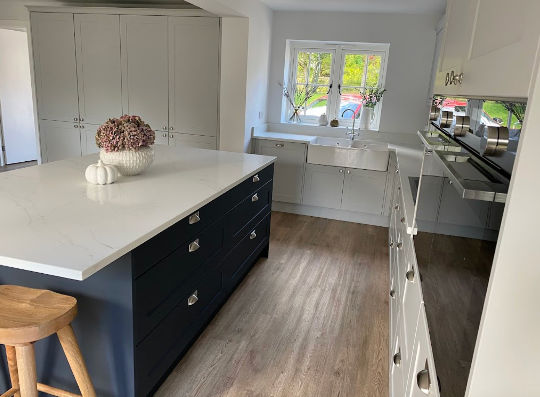 Modern kitchen with a dark blue island, white countertops, tall cabinets, and a farmhouse sink under a bright window.
