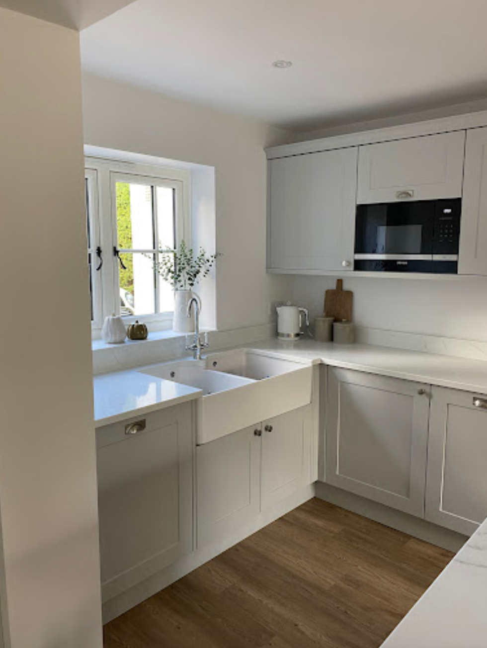 A modern kitchen with light grey cabinets, a white farmhouse sink, white countertops, and wood-look flooring.
