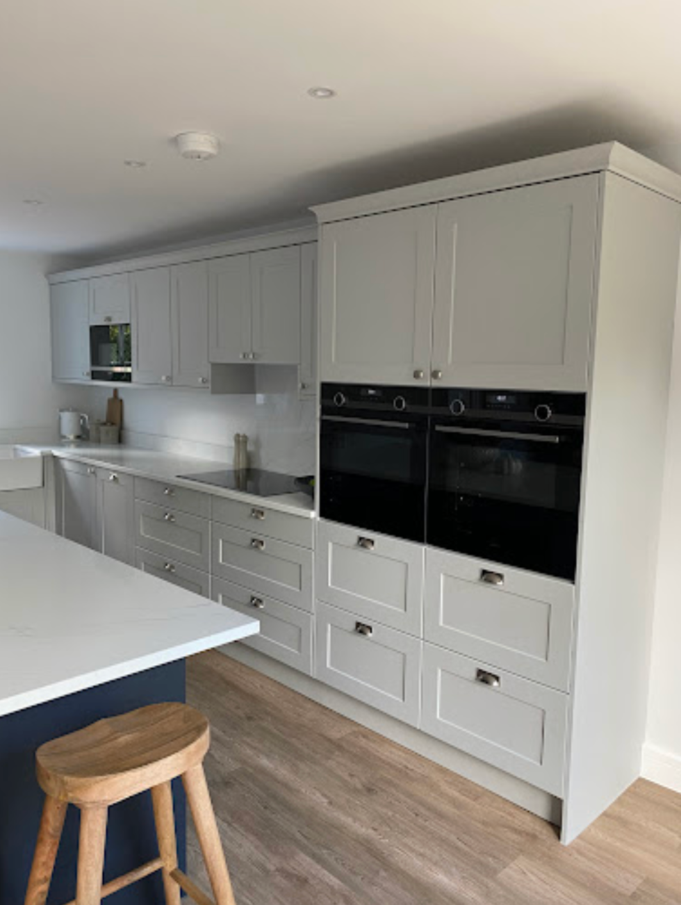 A modern kitchen with light grey cabinets, dual built-in ovens, a white countertop, and a wooden stool in the foreground.