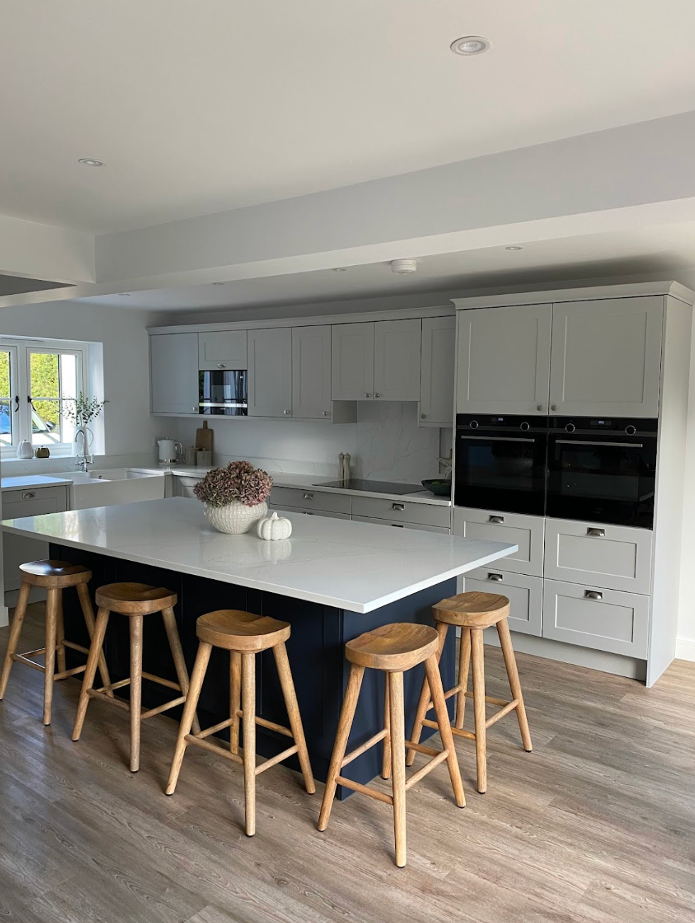 A modern kitchen with light gray cabinetry, a dark blue island, white countertops, and four wooden bar stools.