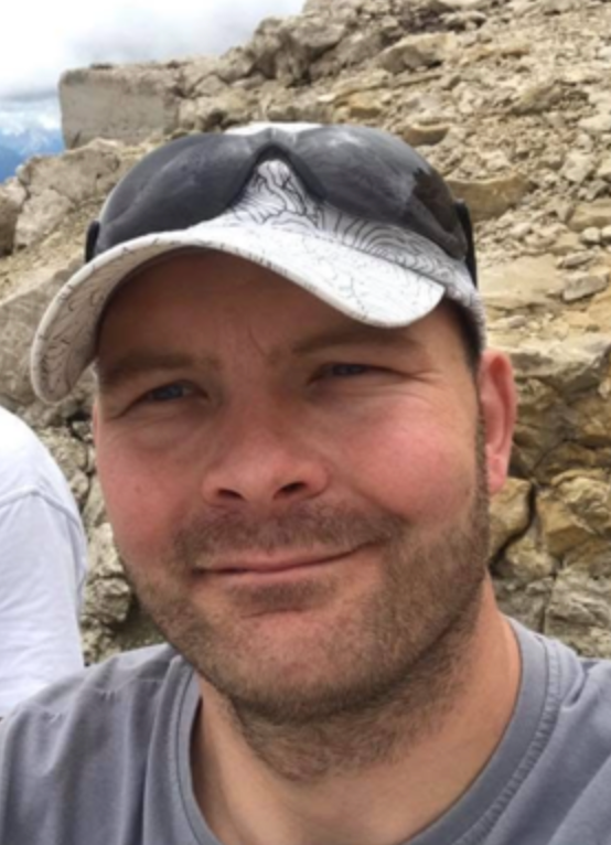 A person wearing a grey topographic print cap and sunglasses against a background of rocky, mountainous terrain.