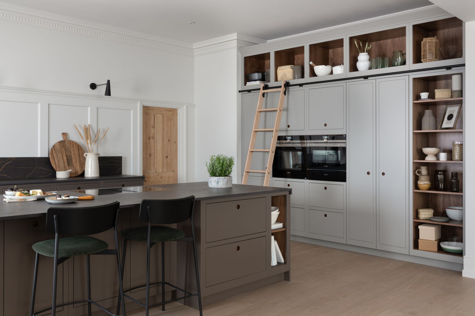 A modern kitchen with a large dark gray island, two black bar stools, and a tall, light gray wall unit with a sliding ladder.