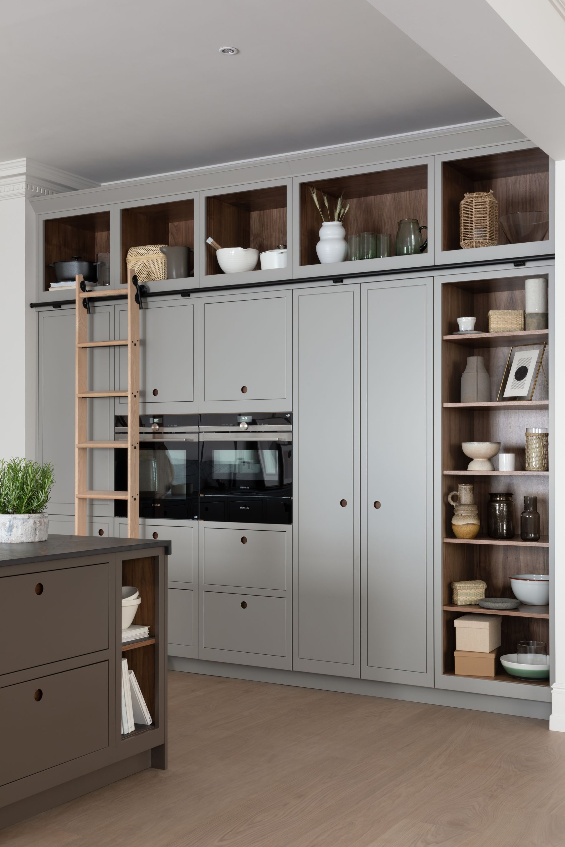 A modern gray kitchen wall featuring built-in ovens, open shelving, and a sliding wooden library-style ladder.