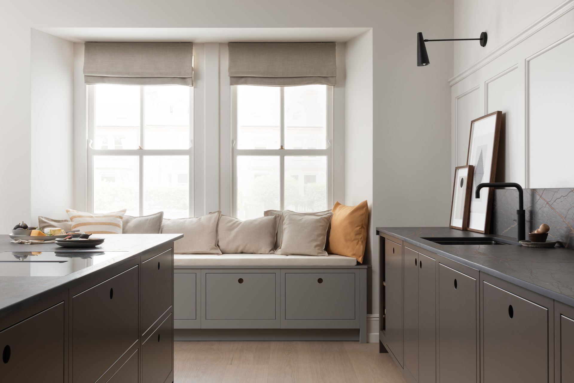A modern kitchen with dark gray cabinets, a window seat with neutral pillows, and white walls.