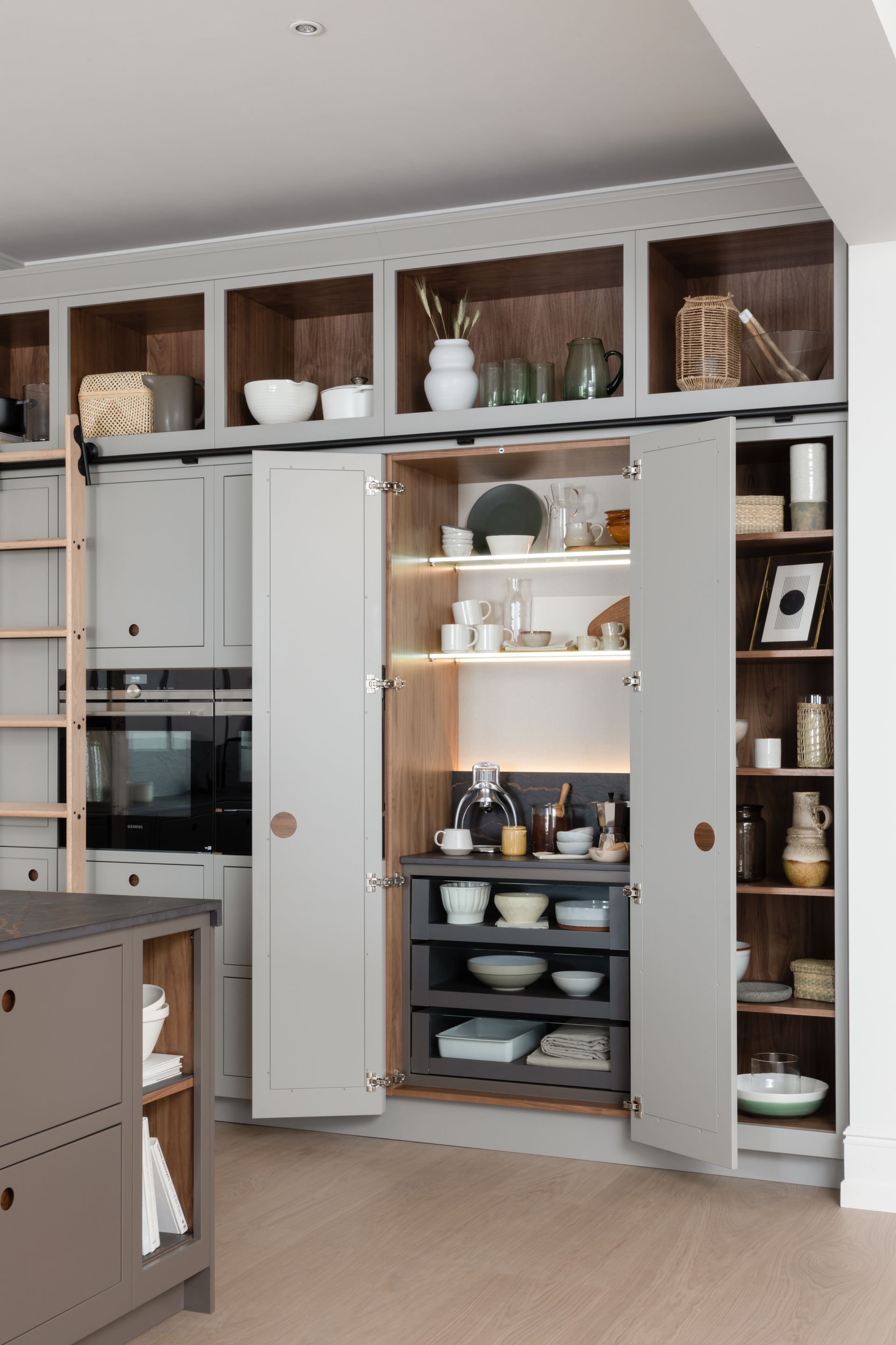 A modern kitchen featuring grey cabinets, open shelving, a built-in coffee station, and a sliding ladder on a rail.