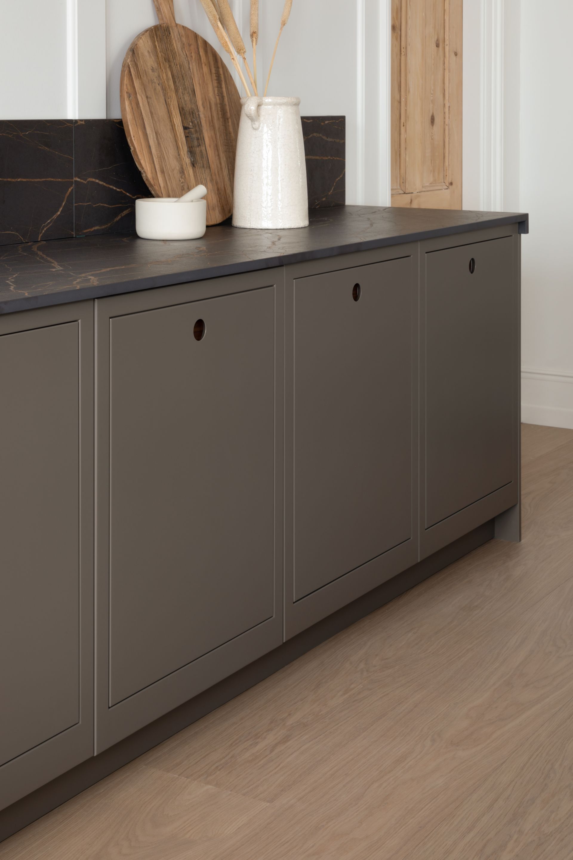 A dark countertop with decor sits atop olive-green cabinets with simple round pulls in a minimalist kitchen setting.