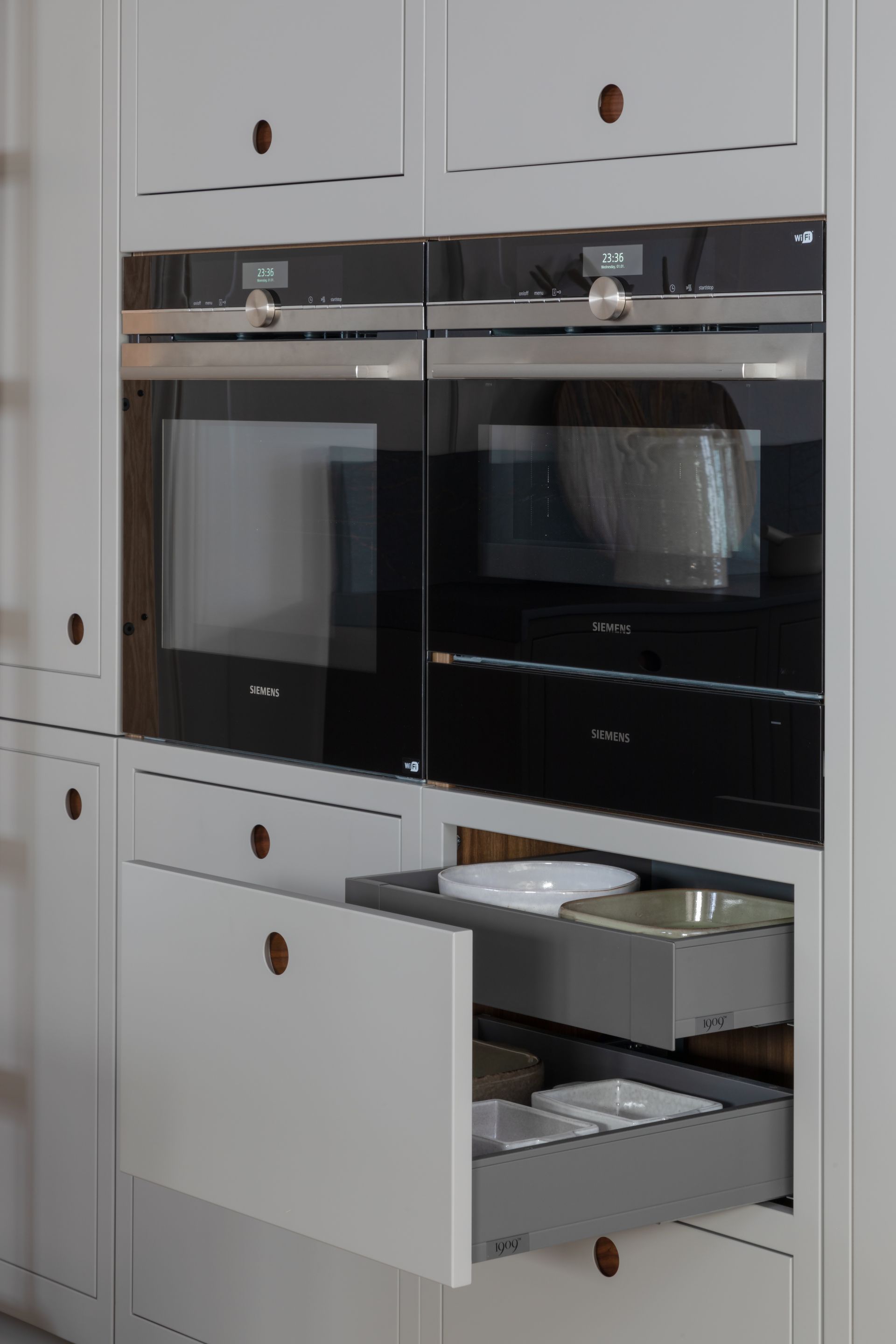 A white cabinet unit featuring two built-in ovens stacked above an open, two-tiered drawer filled with kitchenware.
