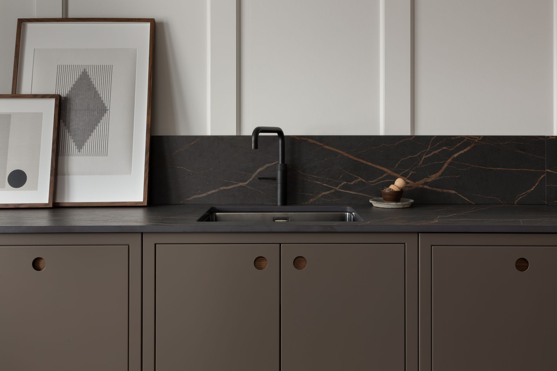A minimalist kitchen counter with dark brown cabinets, a black marble backsplash, a matte black faucet, and framed art.