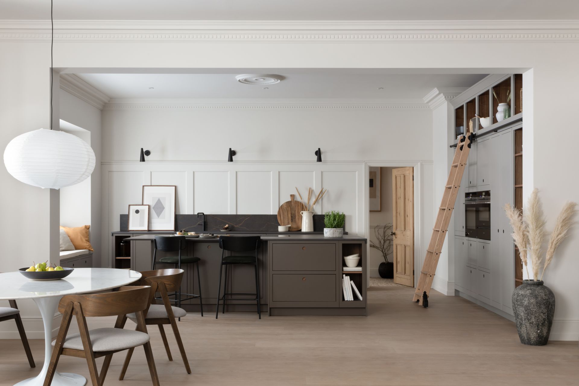Modern kitchen with white walls, a dark grey island, bar stools, a round dining table, and a sliding ladder on cabinets.