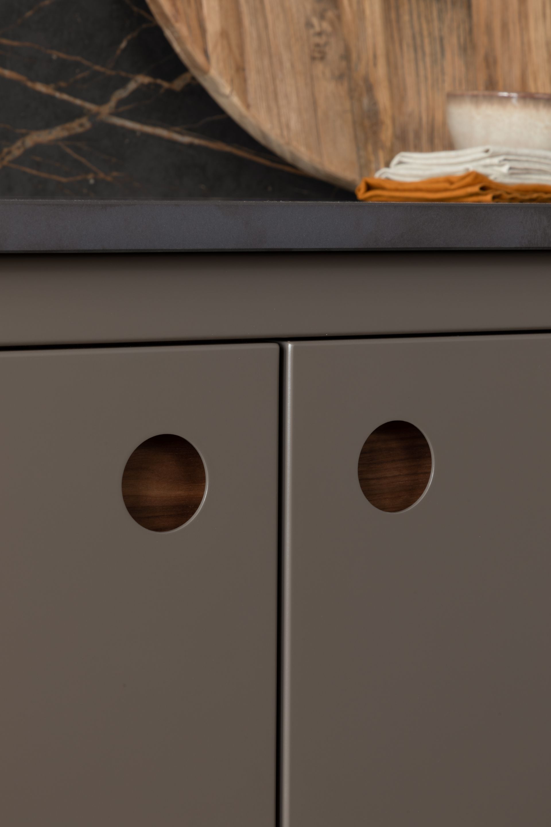 Close-up of matte grey kitchen cabinet doors with circular, dark wood recessed handle pulls beneath a stone countertop.