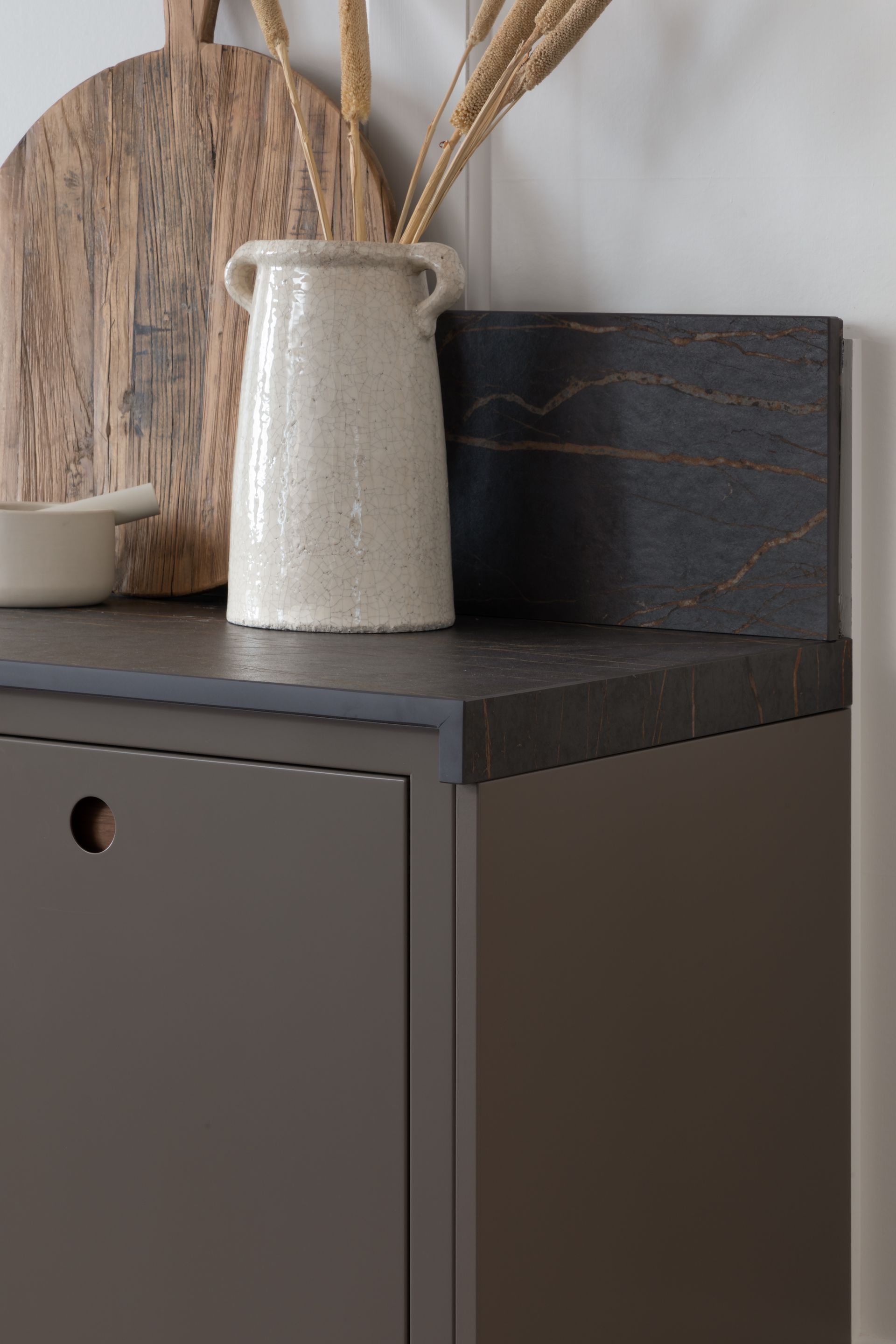 A white ceramic vase with dried grass, a wooden cutting board, and a small bowl sit on a dark, textured kitchen counter.