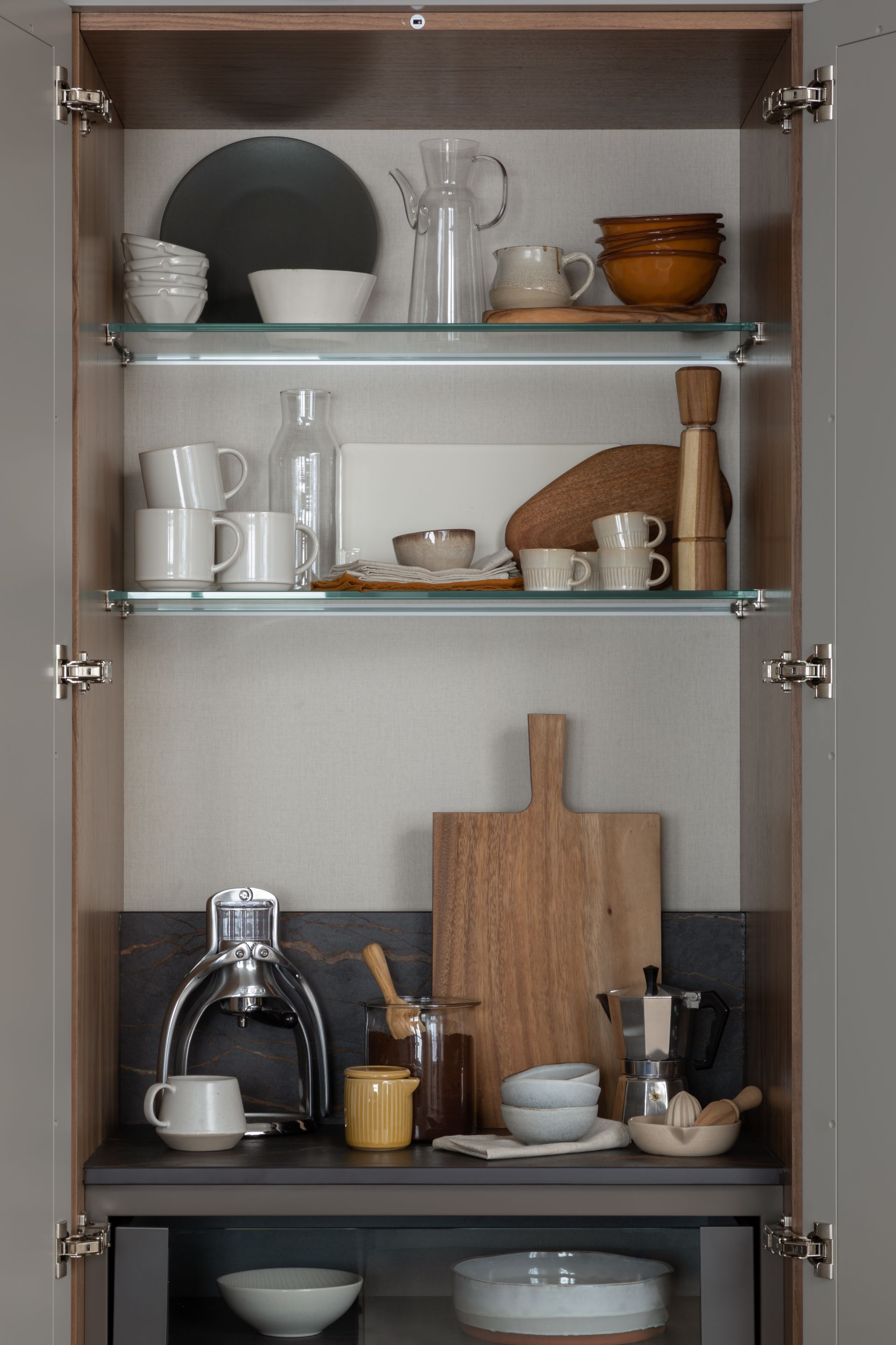A kitchen cabinet with glass shelves holding dishes, glassware, and kitchen tools, with a lower counter for coffee gear.