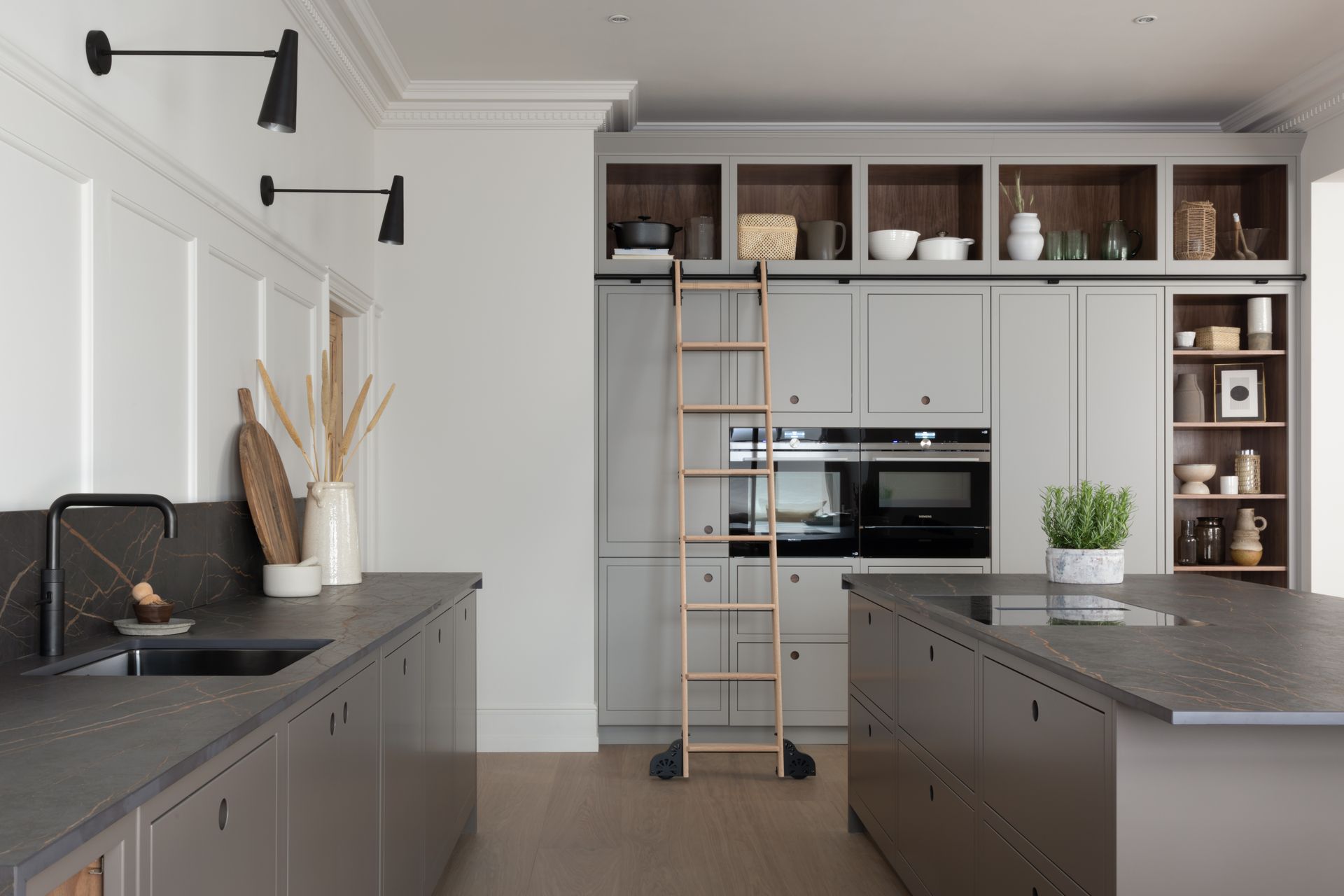 A modern kitchen with grey cabinetry, a black island, a sliding library ladder, and open shelving against white walls.