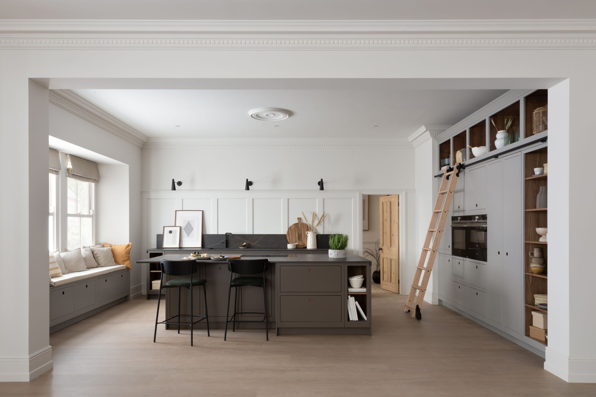 Modern kitchen with a dark grey island, white cabinets, a wooden rolling ladder, and a window seat in a white room.