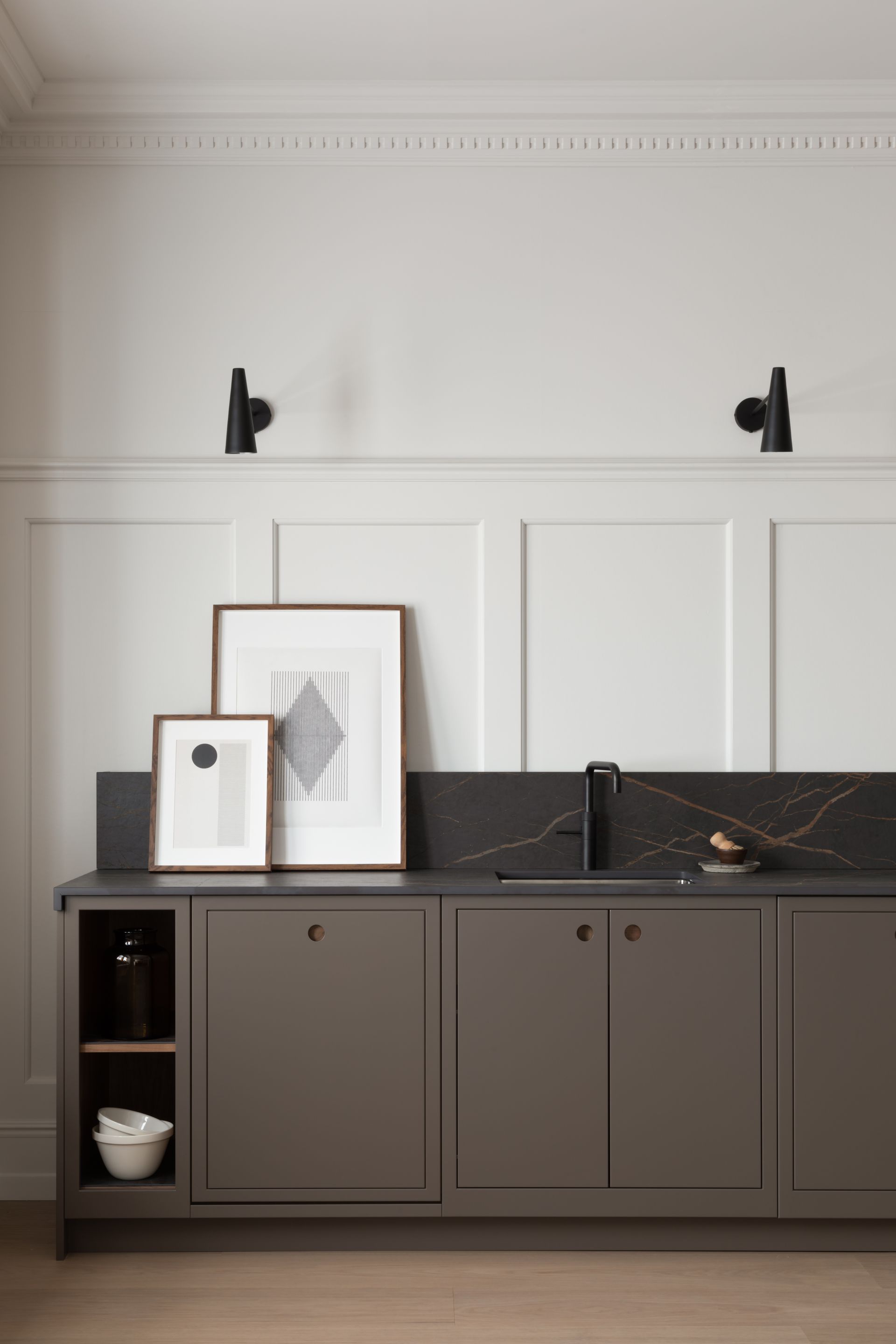 A kitchen counter with dark grey cabinets, a black marble backsplash, and framed artwork against a white paneled wall.