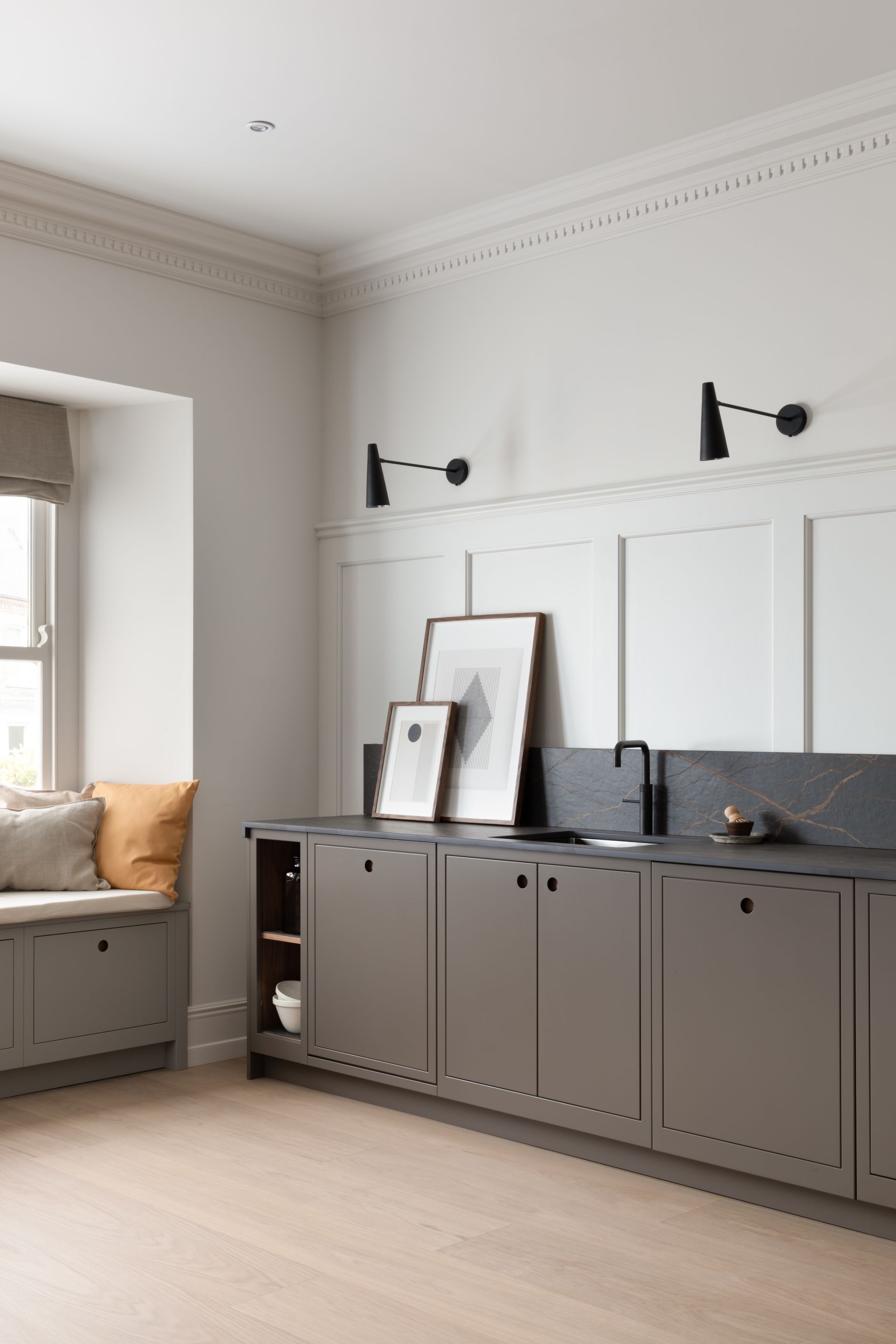 A sleek kitchen featuring taupe cabinets, black countertops, framed artwork, wall sconces, and a window seat with cushions.