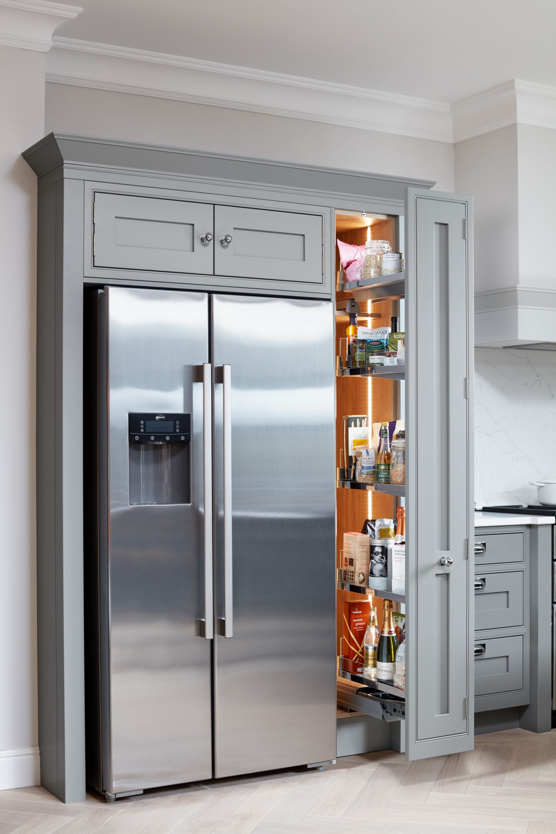 Stainless steel refrigerator beside a lit pull-out pantry cabinet in a kitchen with grey custom cabinetry and molding.