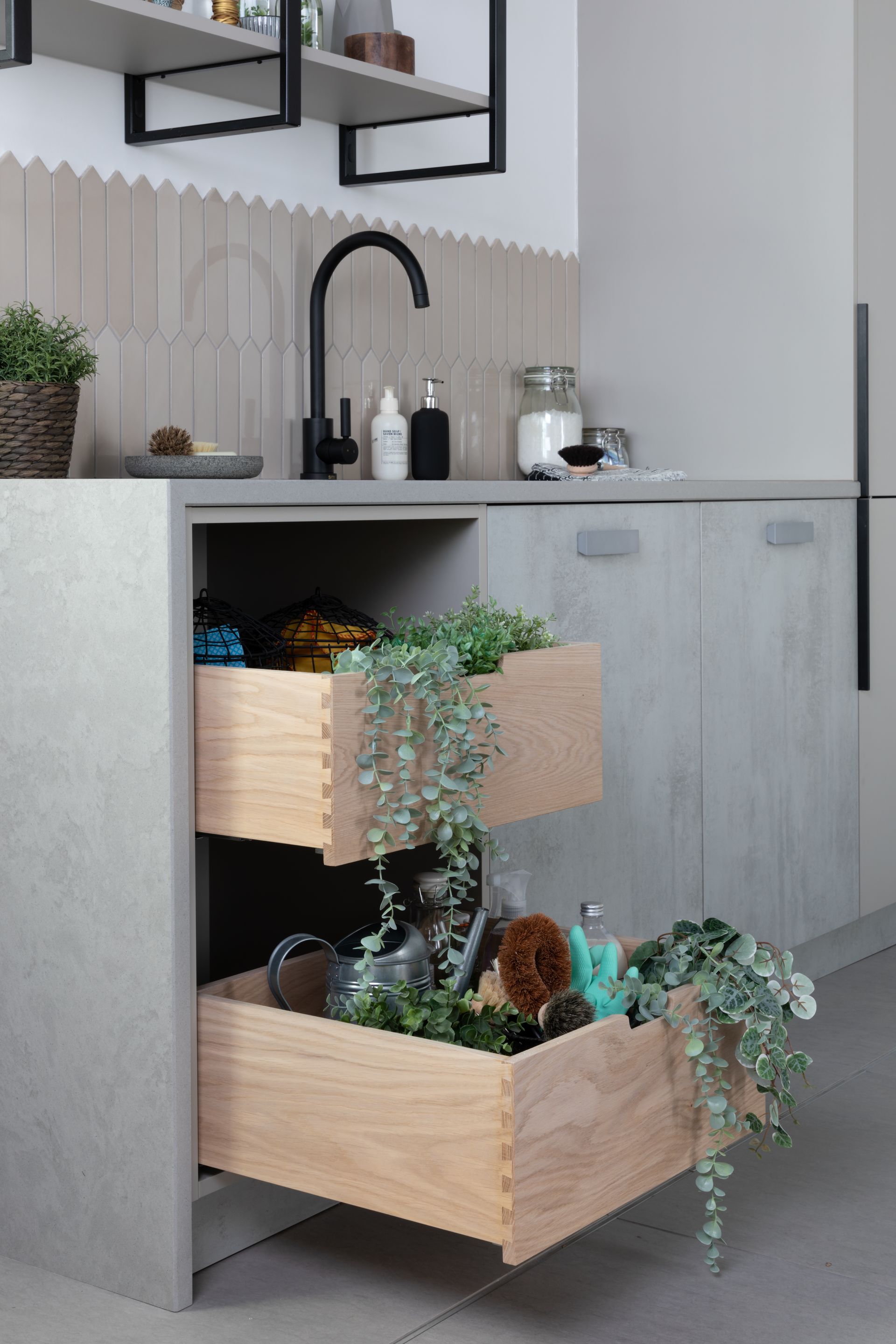 A kitchen cabinet with two open wooden drawers filled with various plants and cleaning supplies under a sink.