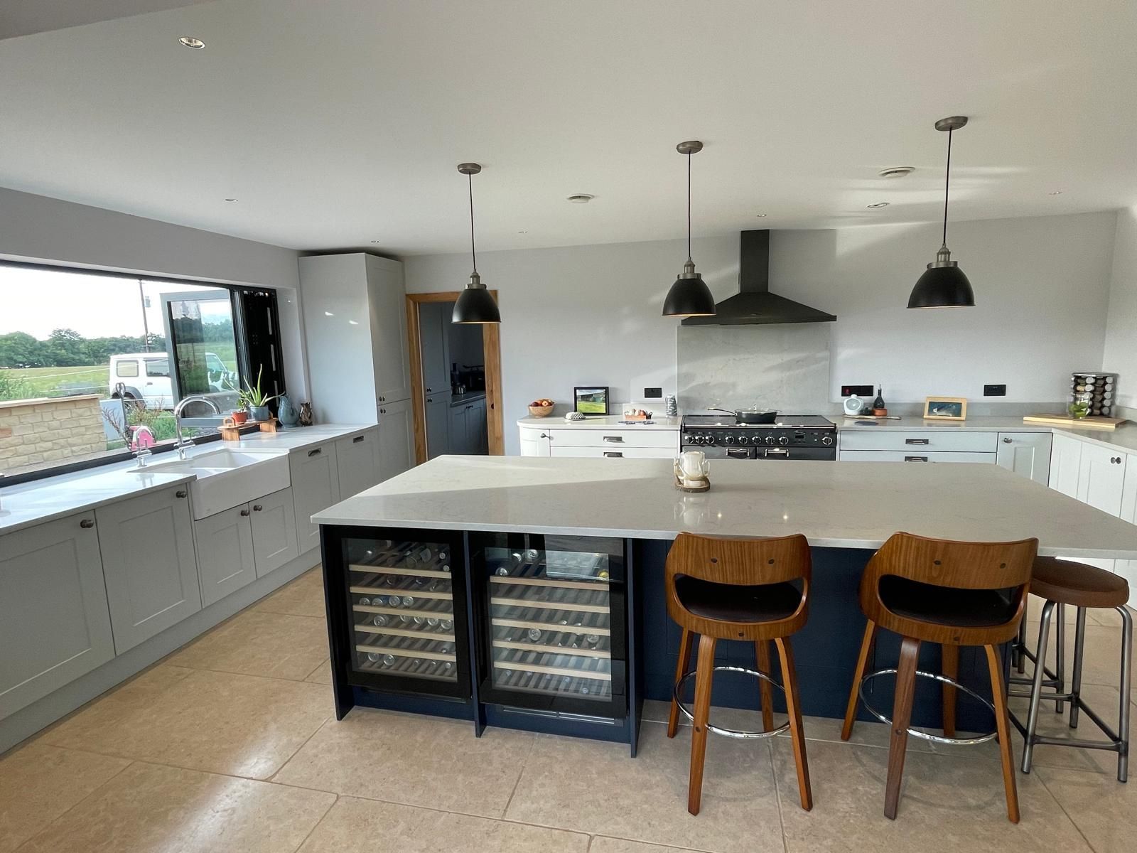 A modern, open-plan kitchen with grey cabinets, a white island with wine fridges, wooden stools, and large windows.