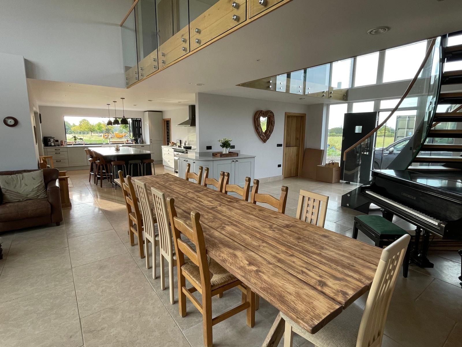 A long wooden dining table with chairs in a modern, open-plan home featuring a grand staircase and kitchen in the back.