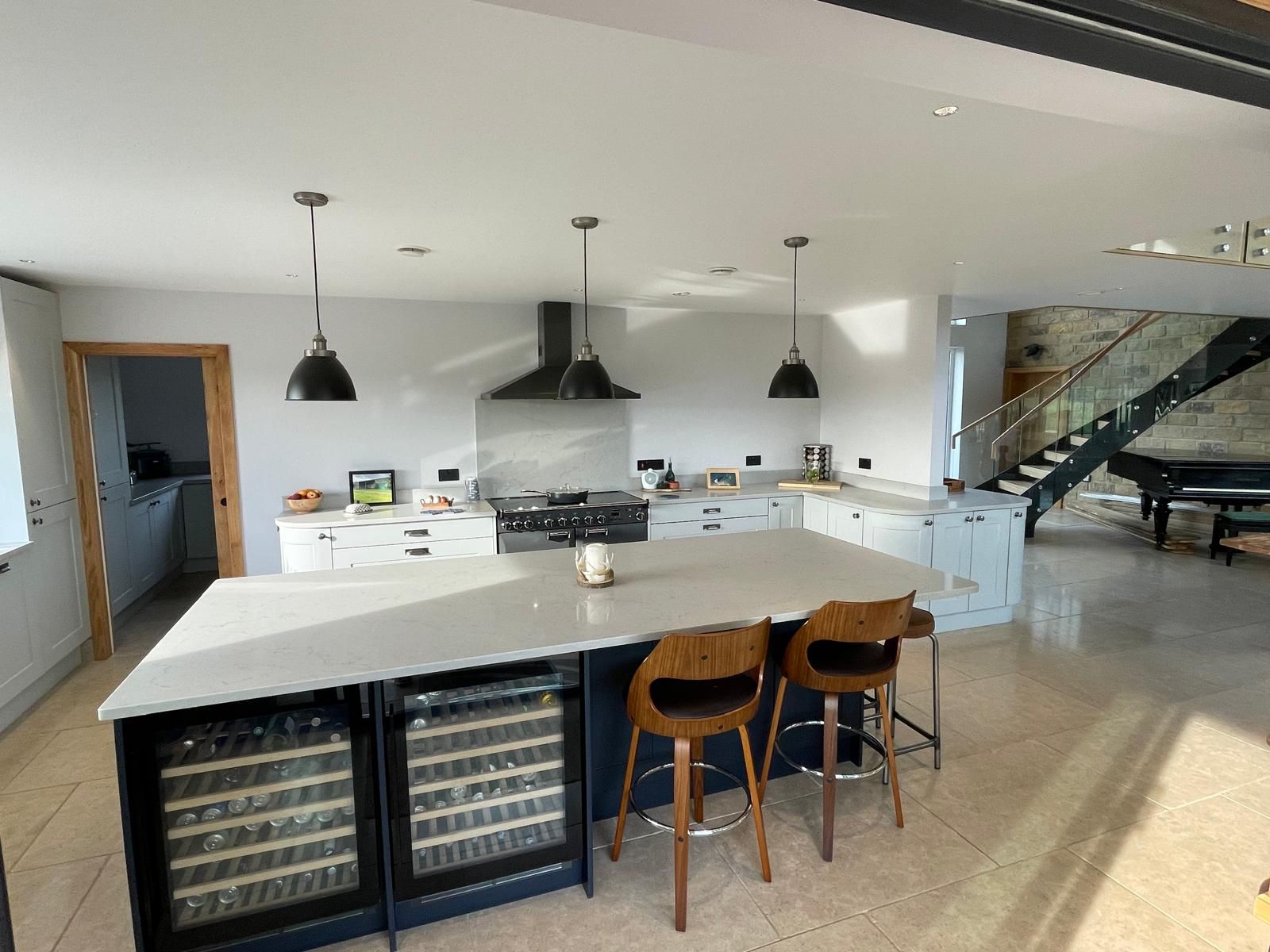 A modern kitchen with a large gray island, two wine fridges, three wooden bar stools, and a staircase in the background.