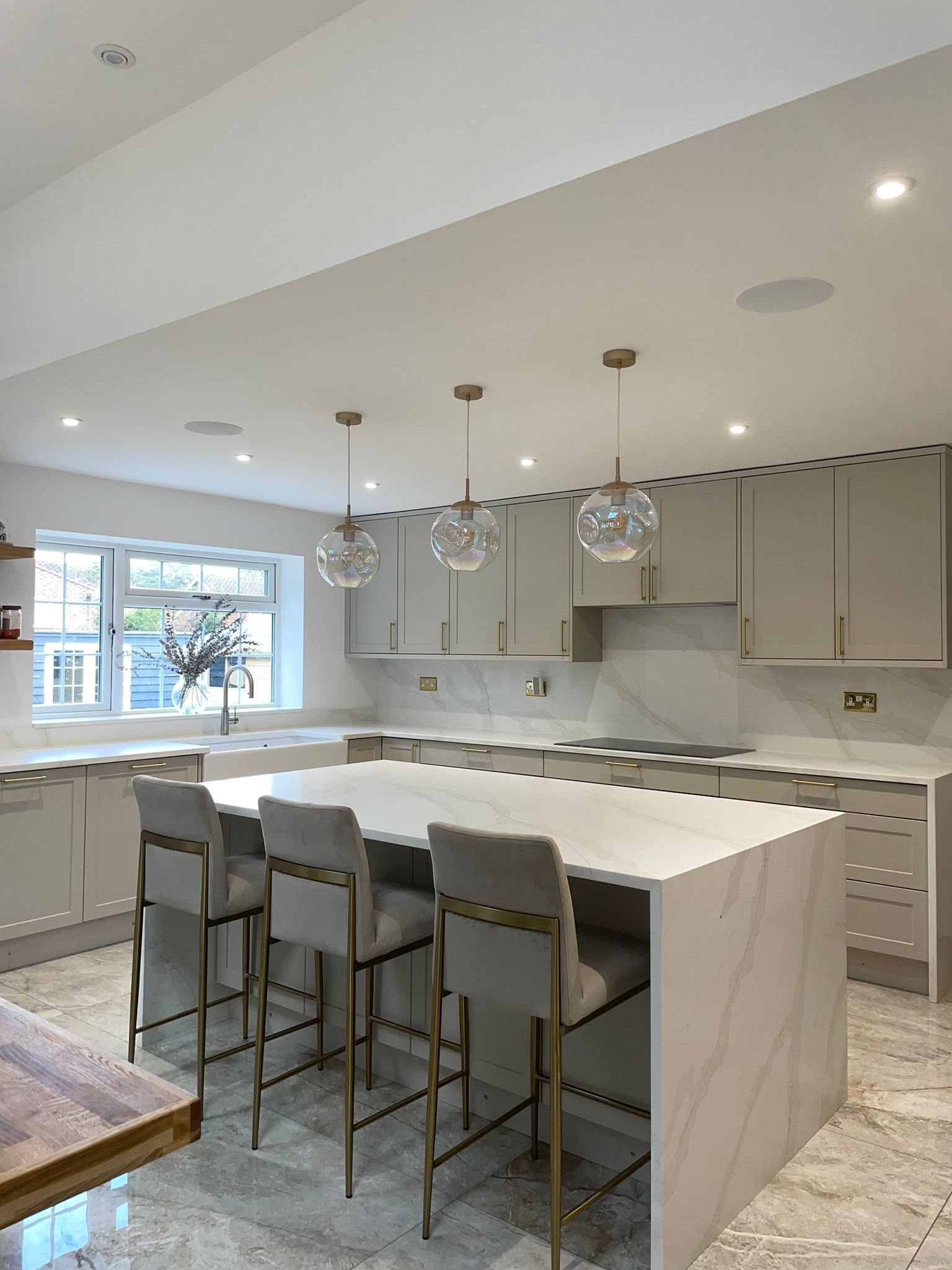 A modern kitchen with light gray cabinetry, marble island countertops, three pendant lights, and gray bar stools.