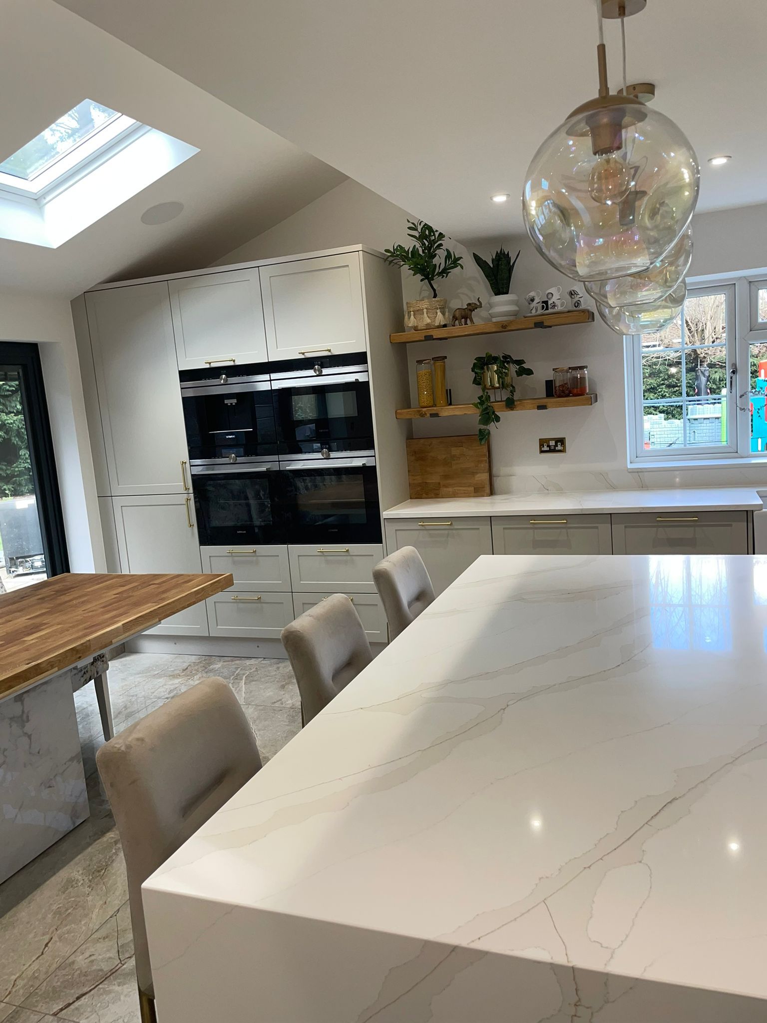 A bright modern kitchen with a large white marble-topped island, beige chairs, and light wood shelving under a skylight.