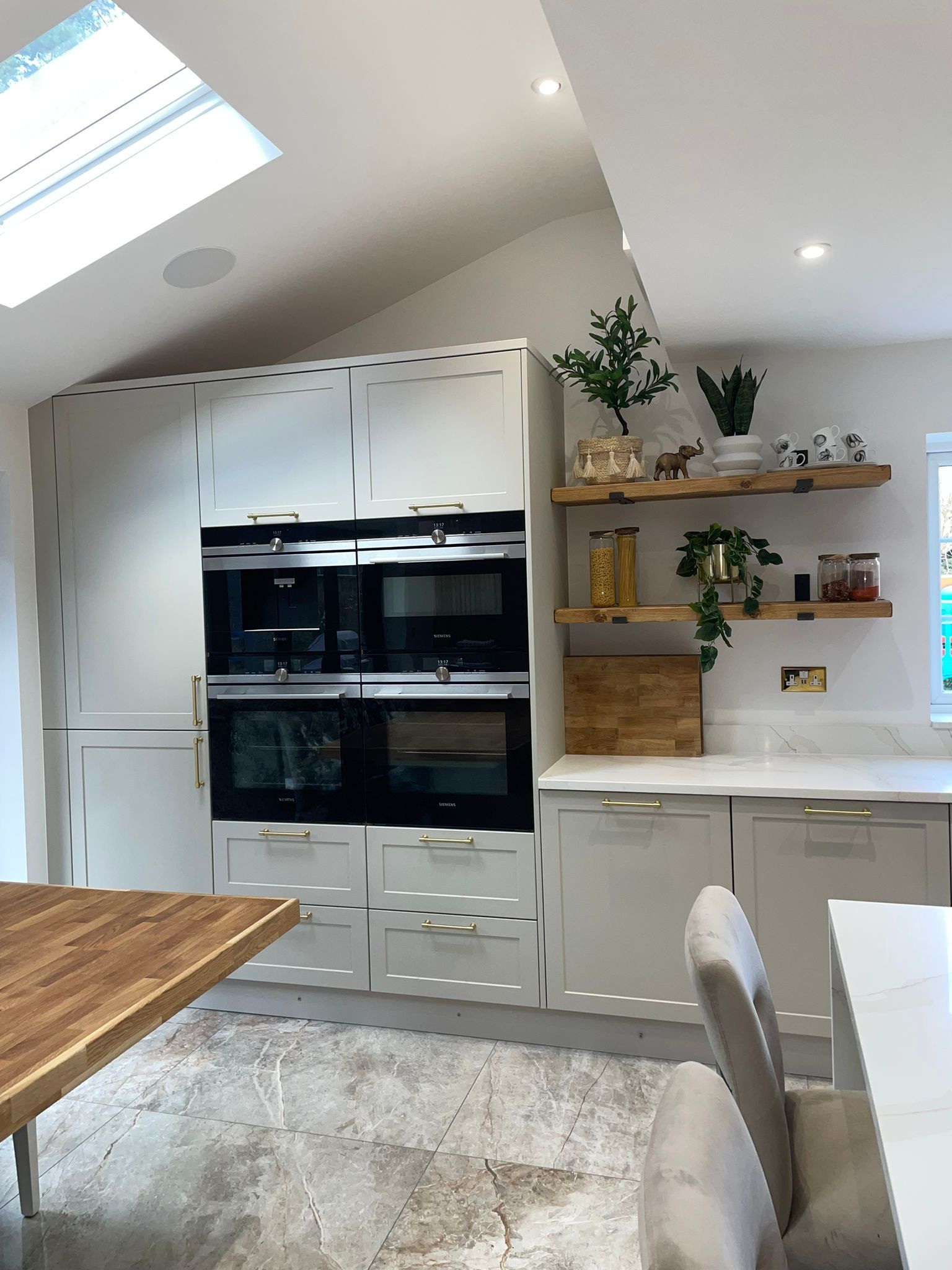 Modern kitchen with a built-in double oven, cream cabinetry, open wooden shelving with plants, and a tiled floor.
