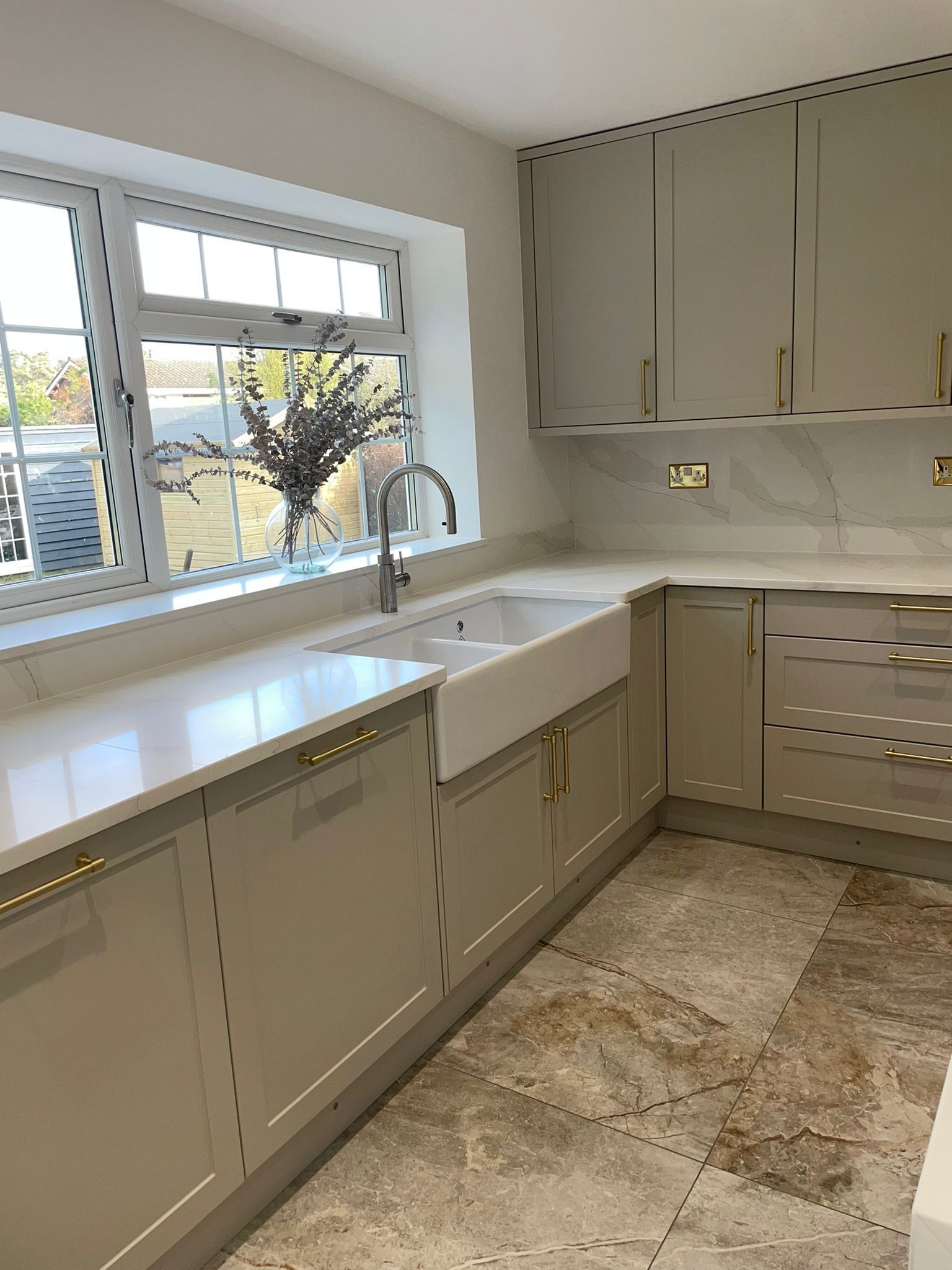 A modern kitchen featuring light taupe cabinets, gold hardware, a white farmhouse sink, and marble countertops.