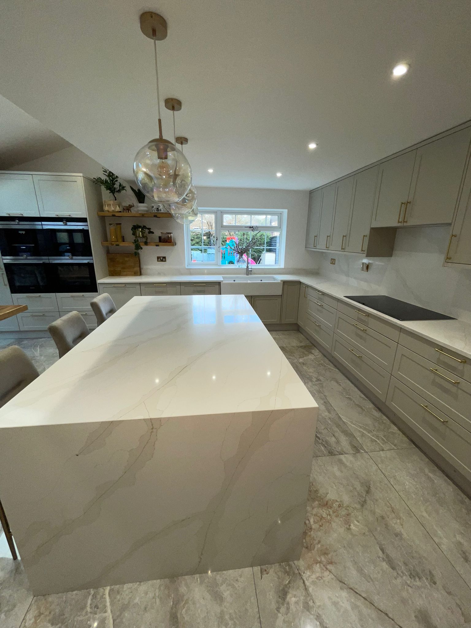 A modern kitchen featuring a large white marble-topped island, light grey cabinets, and stone tile flooring.