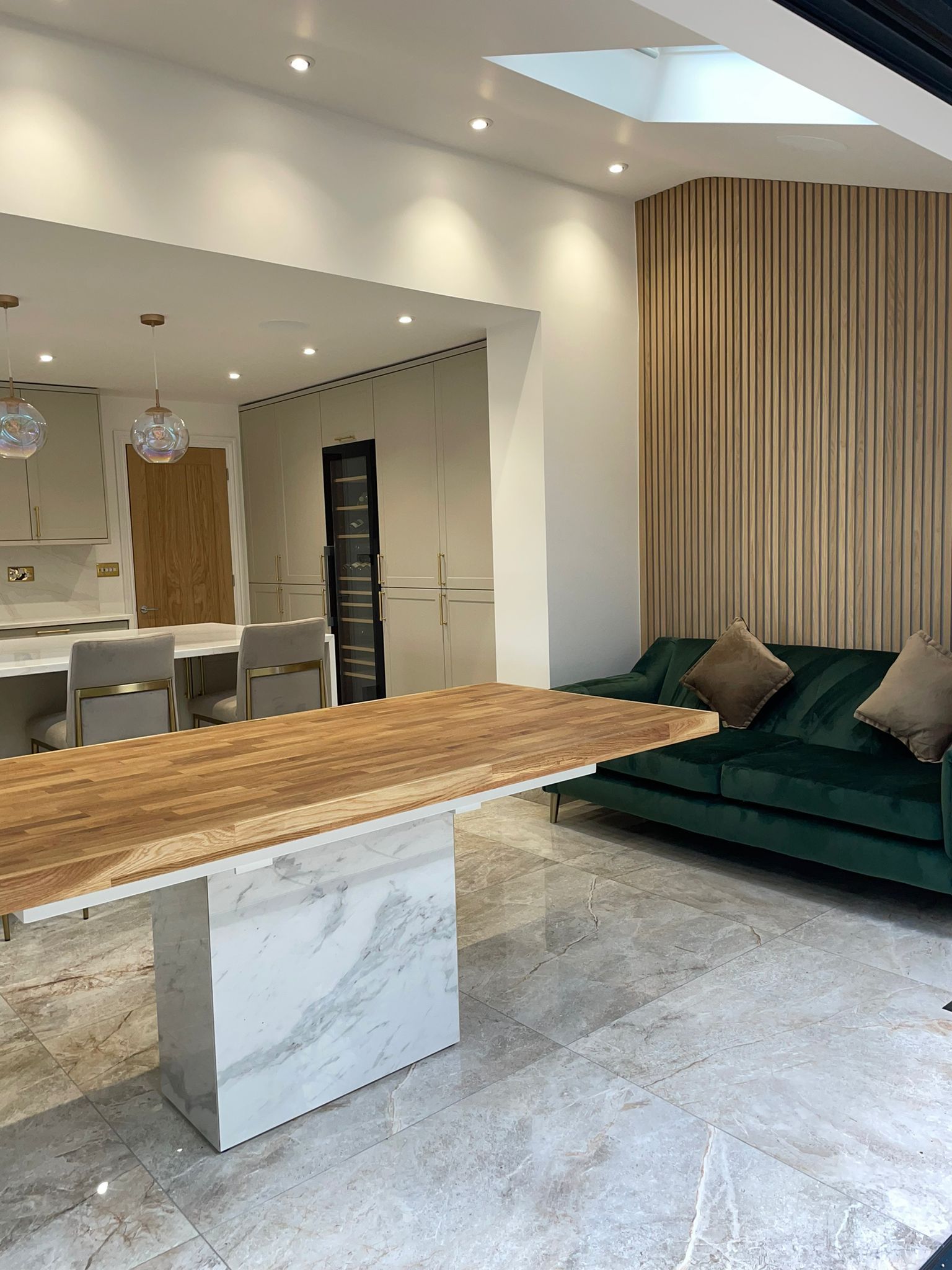 A modern room featuring a marble-based wood table in the foreground, a deep green velvet sofa, and light wood wall slats.