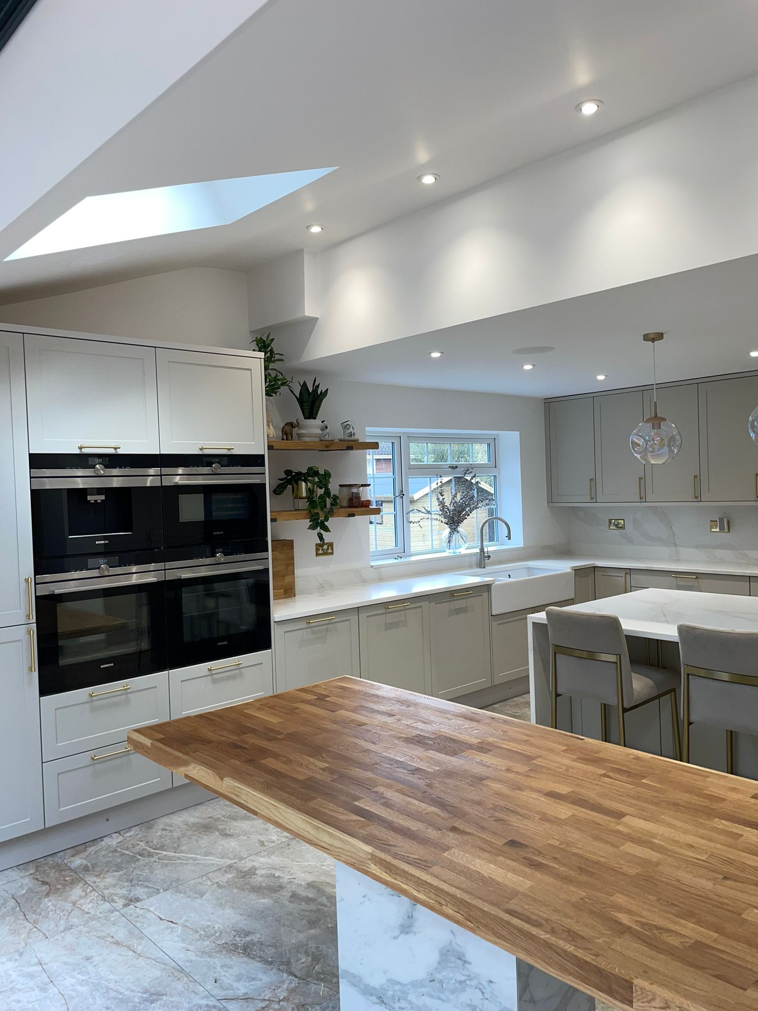 Modern kitchen with white cabinets, wood-topped island, marble flooring, and an oven stack under a pitched skylight ceiling.