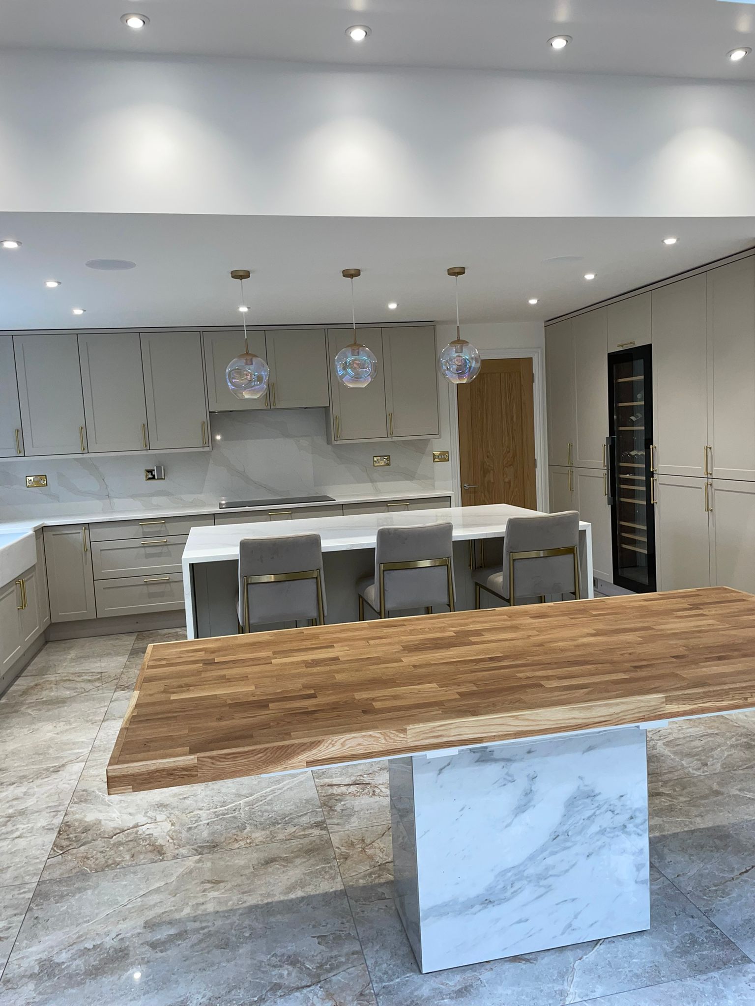 A modern kitchen with light-colored cabinets, a marble-topped island with chairs, and a large wooden breakfast bar.