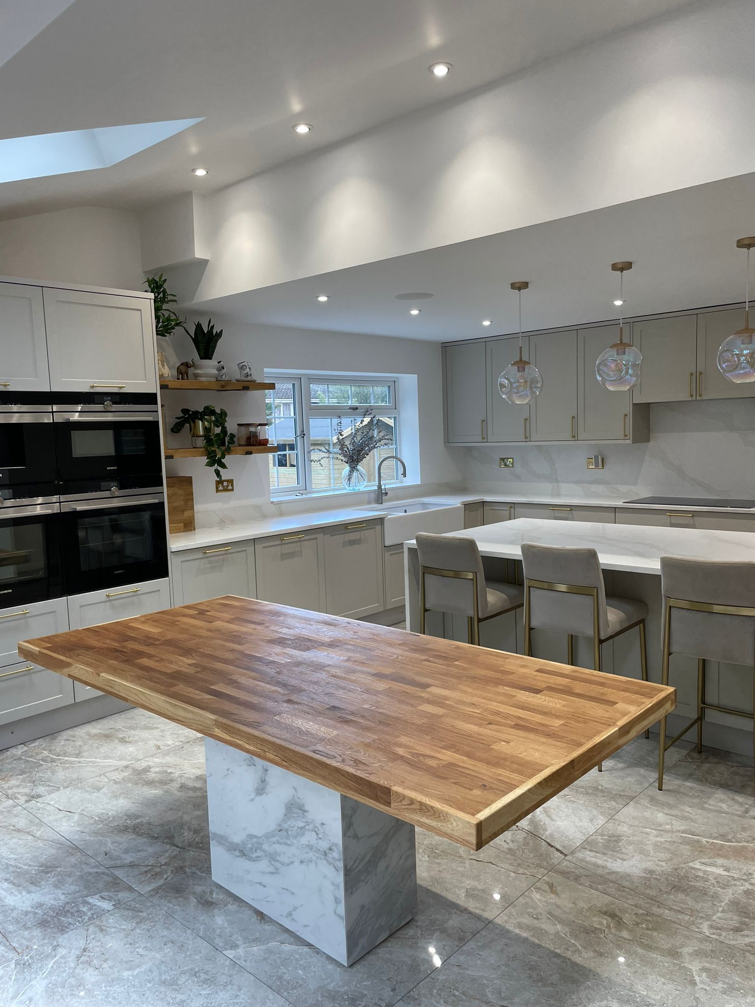 A modern kitchen featuring a marble-based wood dining table, white cabinets, and marble flooring under recessed lighting.