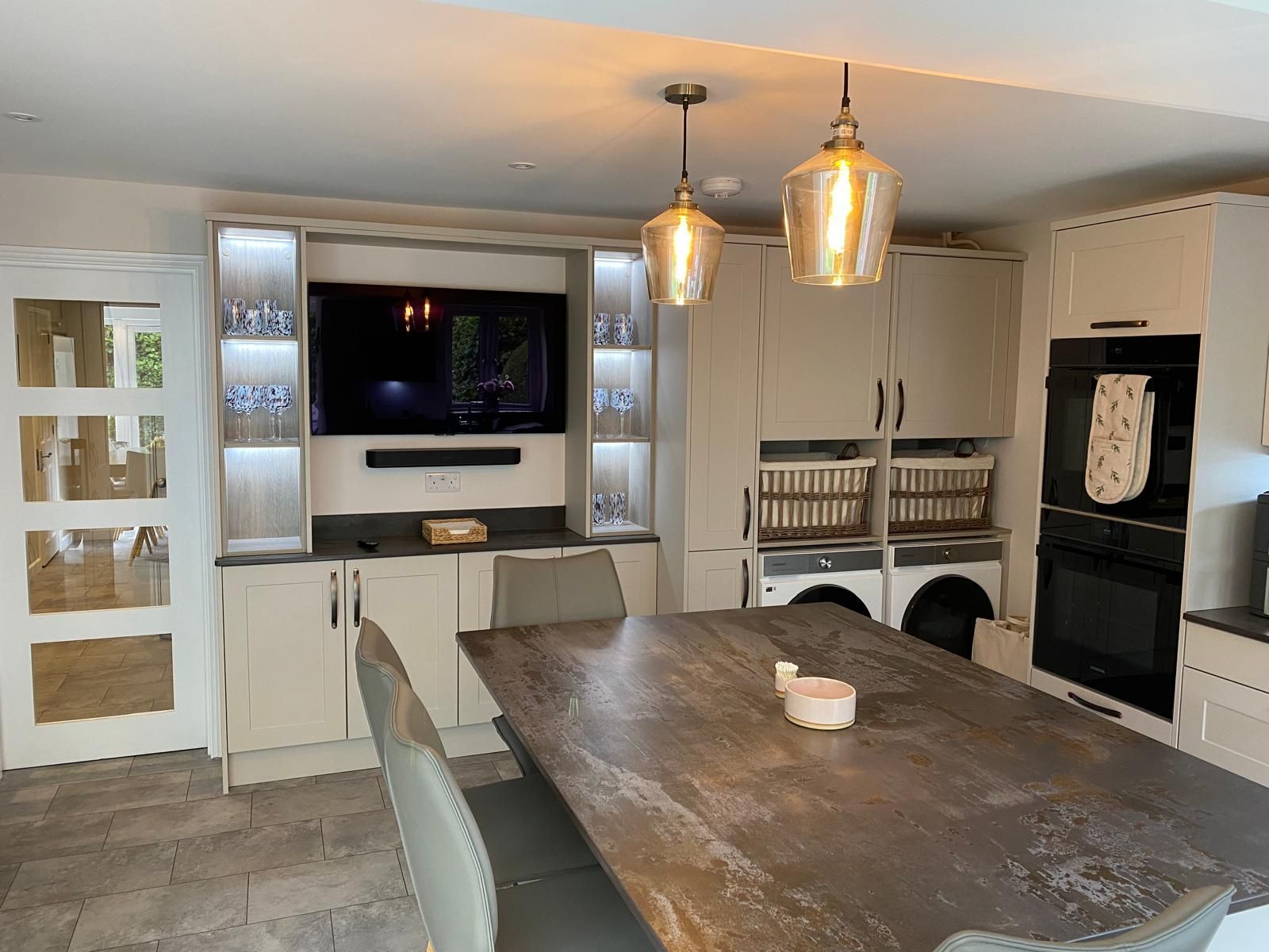 A dining area featuring a large, dark stone table, neutral cabinetry, a wall-mounted TV, and two glass pendant lights.