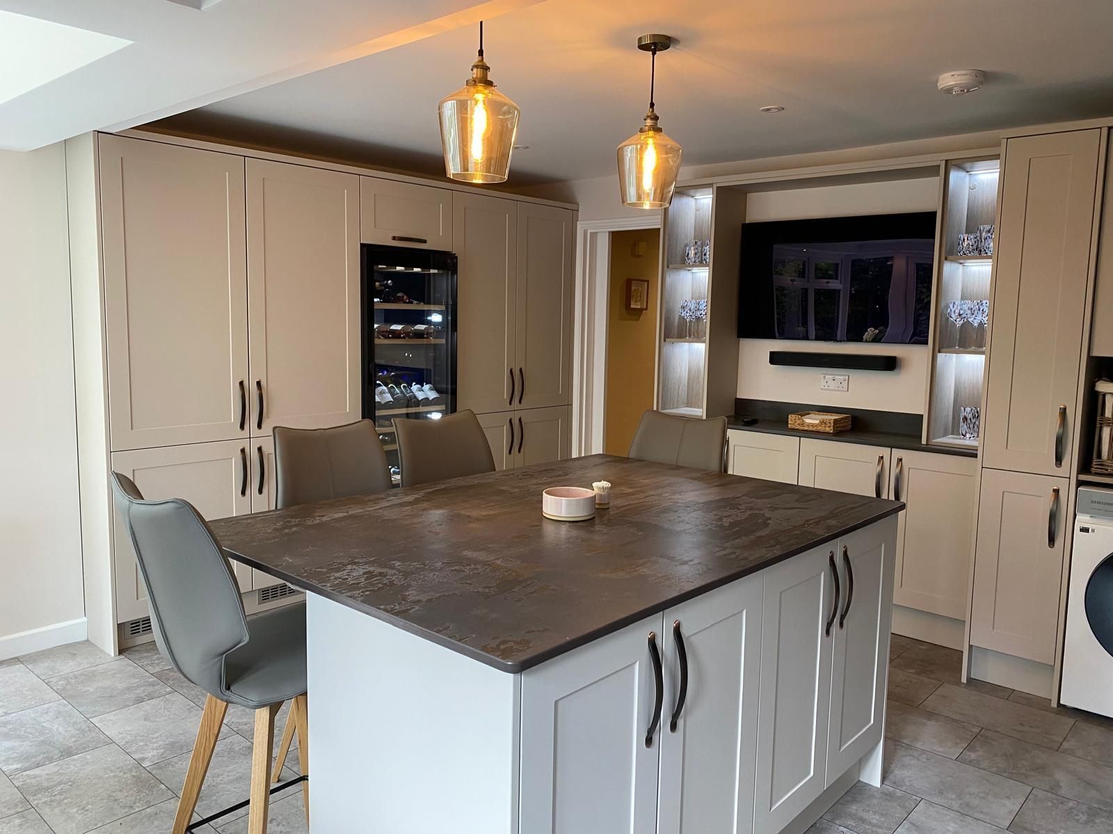 Modern kitchen with a central island, grey chairs, beige cabinets, pendant lighting, and a wall-mounted TV.