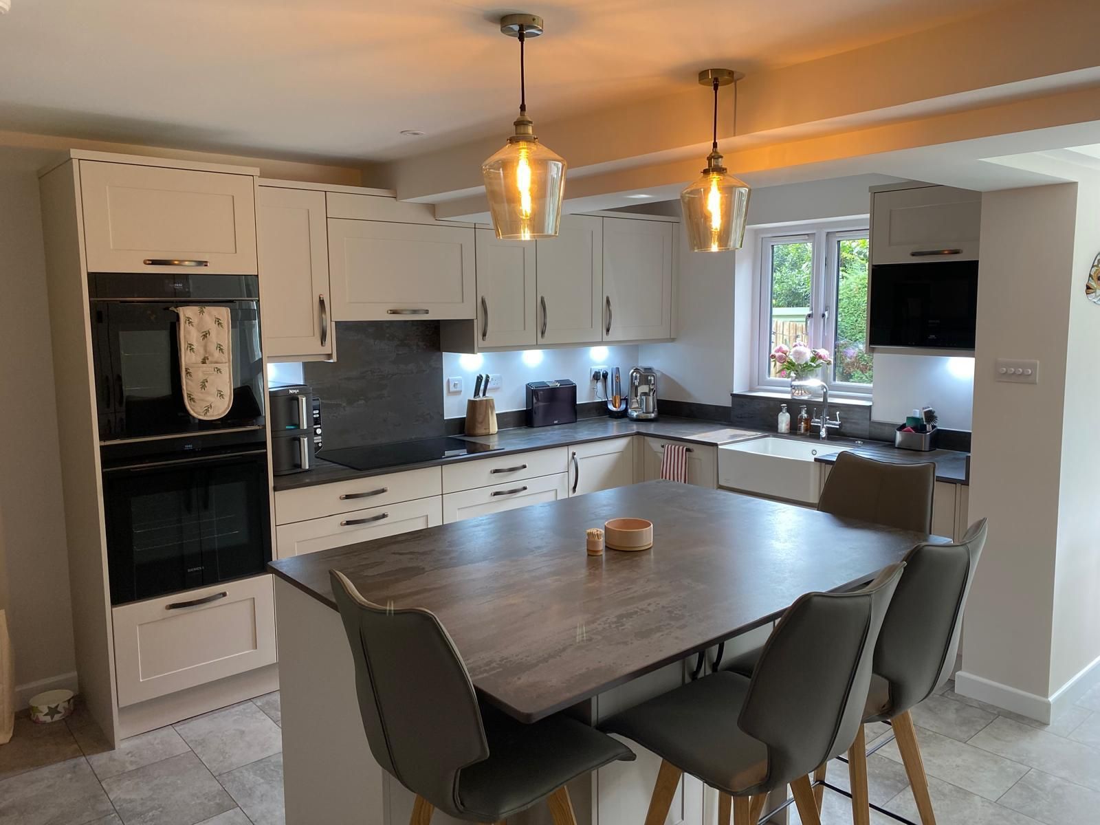 A modern kitchen with cream cabinets, a dark wood island, grey upholstered chairs, and two hanging pendant lights.