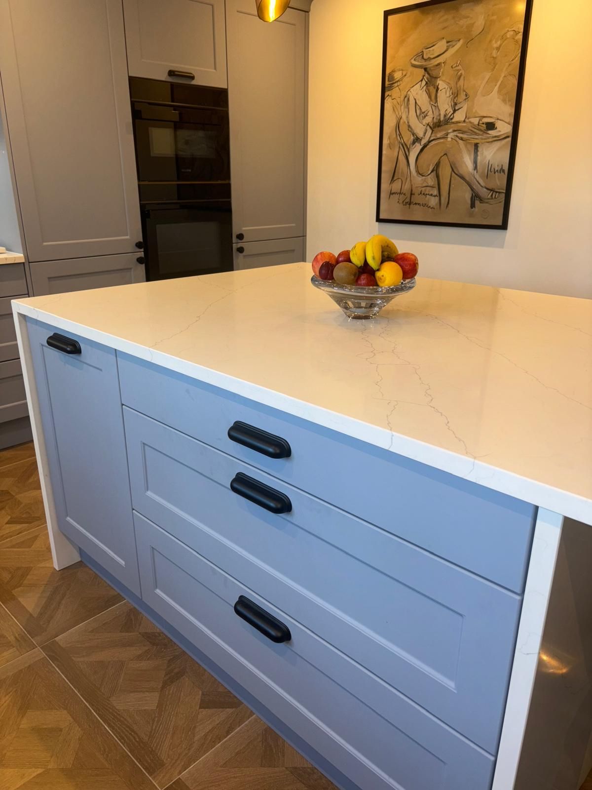 A light blue kitchen island with three drawers, a white stone countertop holding a bowl of fruit, and a framed artwork.