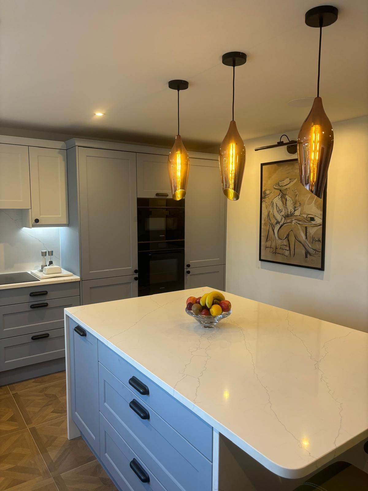 A kitchen island with a white countertop and fruit bowl, set under three amber-glass pendant lights in a modern kitchen.